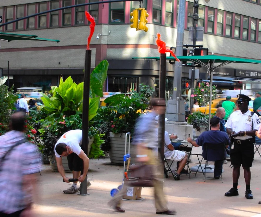 City sidewalk scene with people walking around bright orange figure sculptures on tall poles and sitting, police officer, large green plants, traffic lights, and blurred moving individuals.