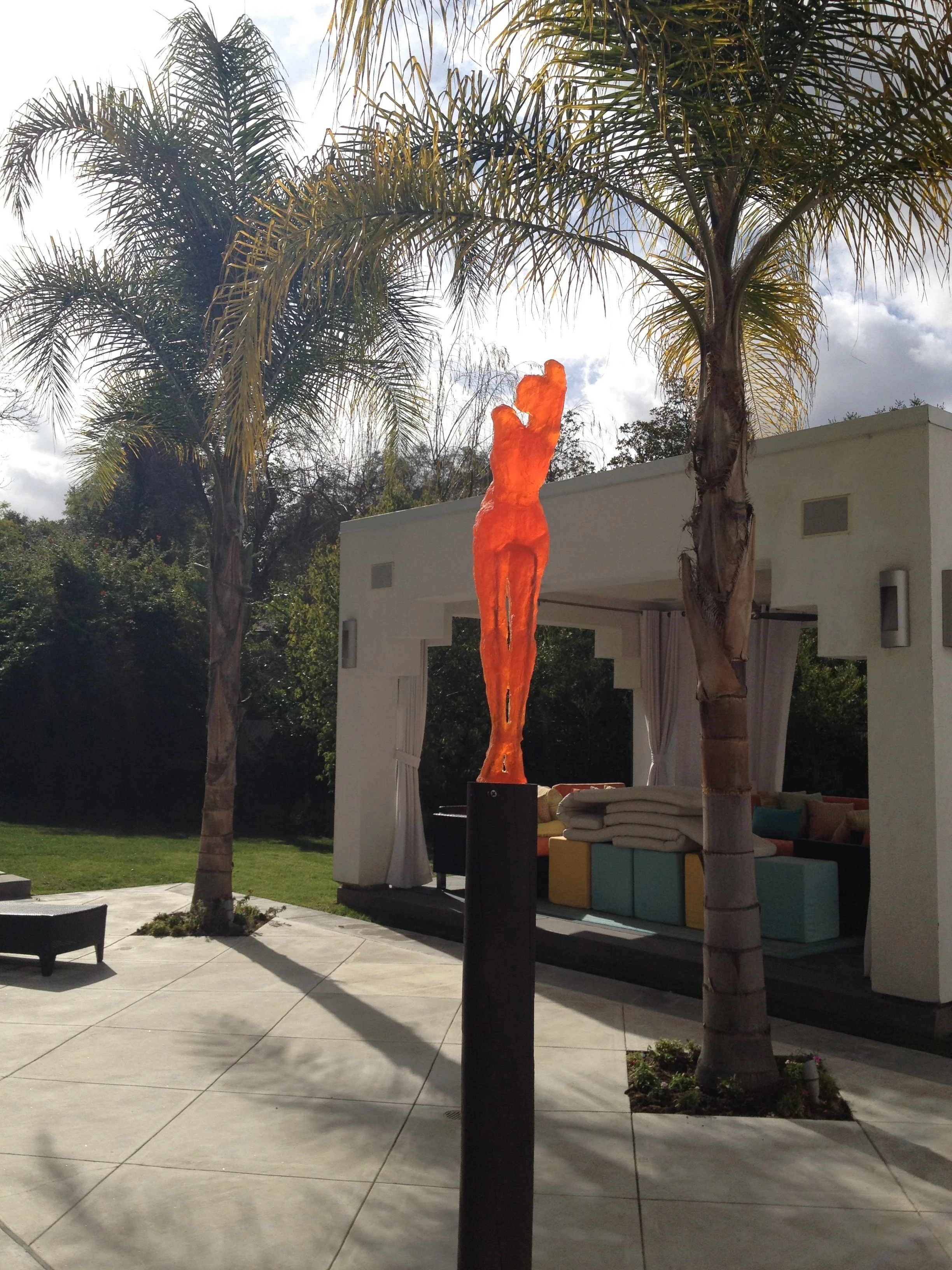Outdoor patio with two palm trees, a white cabana with draped curtains, colorful seating cushions, and a modern sculpture of a person in front of the cabana with the sky in the background.