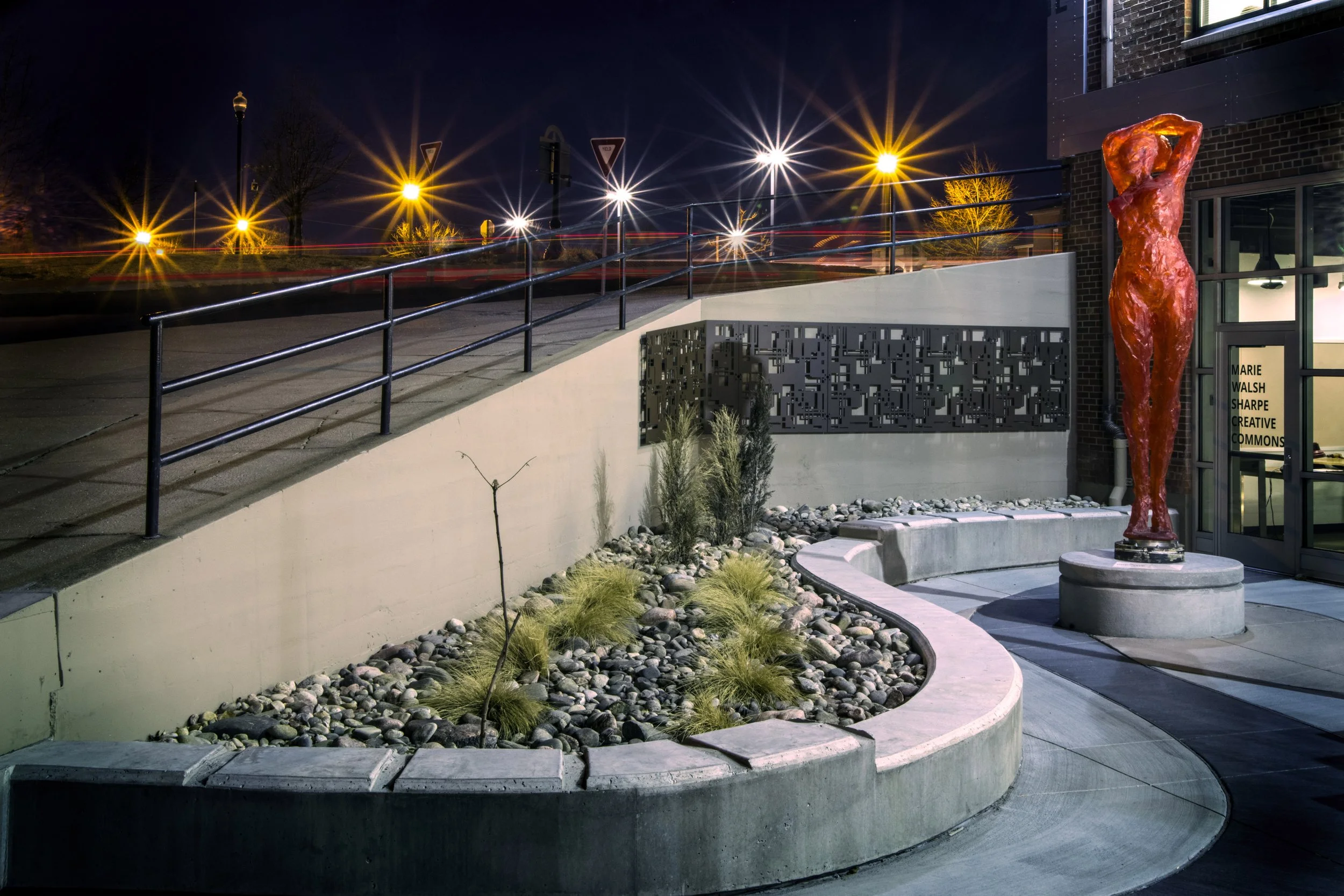 Nighttime scene outside a building with a red abstract human sculpture near the entrance. The scene is illuminated by bright streetlights creating starburst effects, with light trails from passing cars visible in the background. There is a staircase,