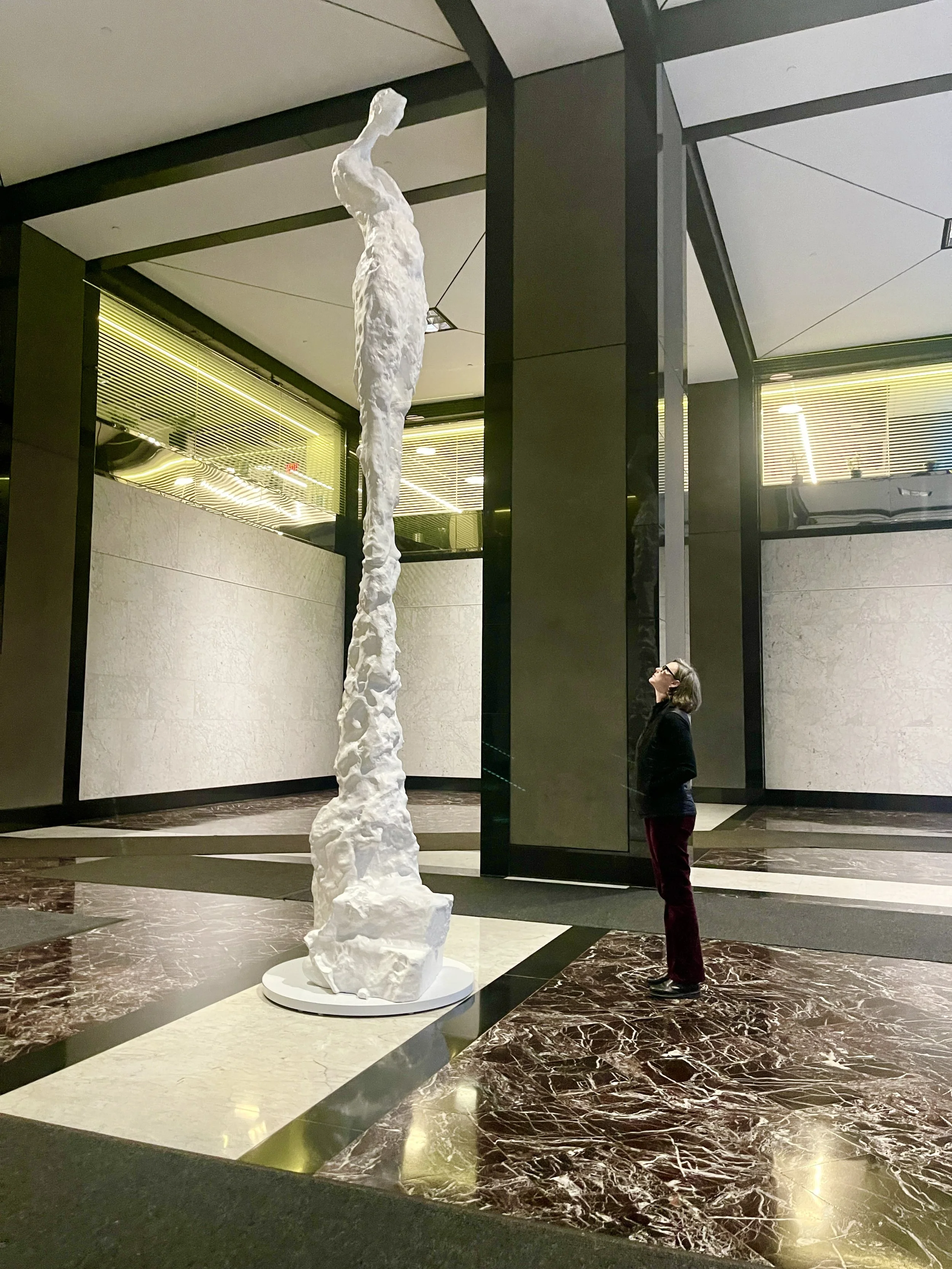 A woman stands on a marble floor looking up at an extremely tall abstract white figurative sculpture inside a modern building with black and cream-colored walls and a ceiling with some light fixtures.