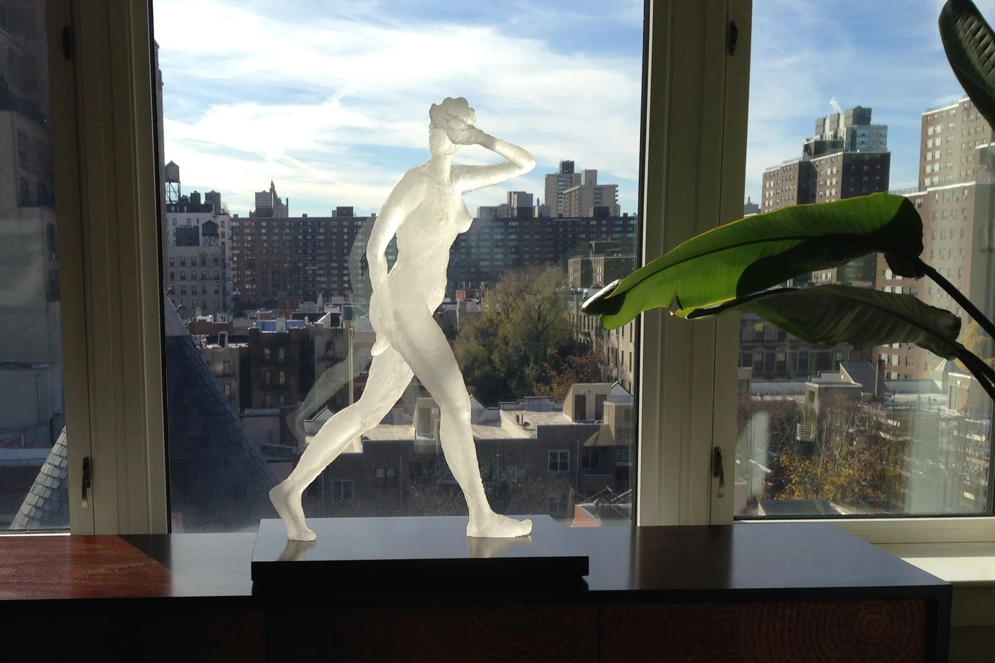 A  sculpture of a woman walking, with one hand on her forehead, positioned on a black base in a window sill. Outside the window, a cityscape with various buildings and a blue sky is visible, along with part of a large green plant.