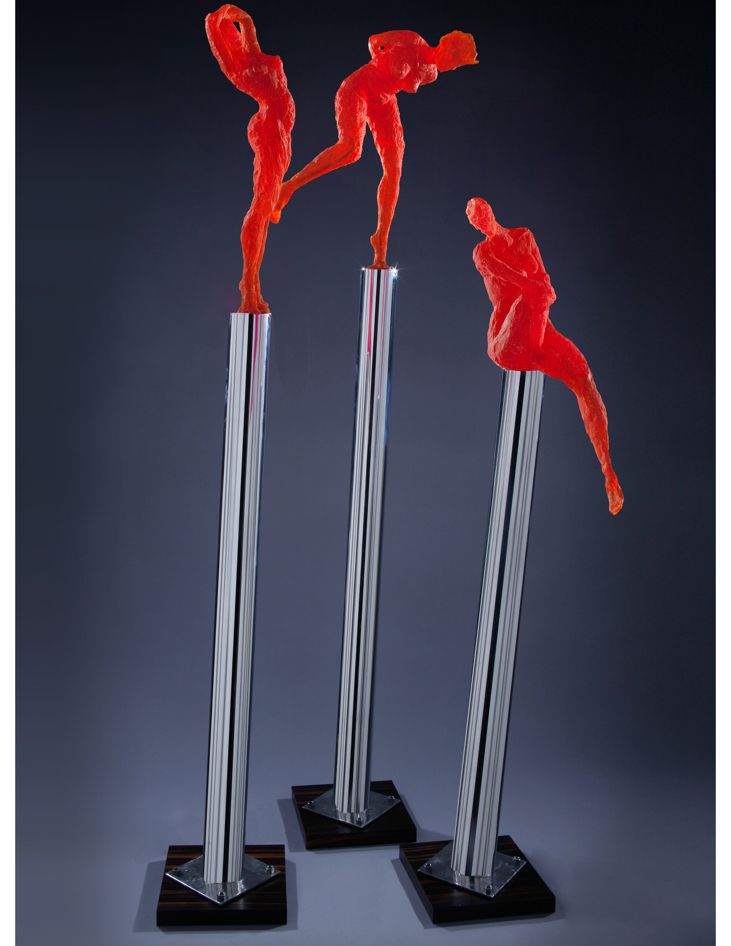 Three modern figure sculptures with red, abstract, textured figures mounted on tall, metallic pedestals with black bases, displayed on a gray surface with a dark background.