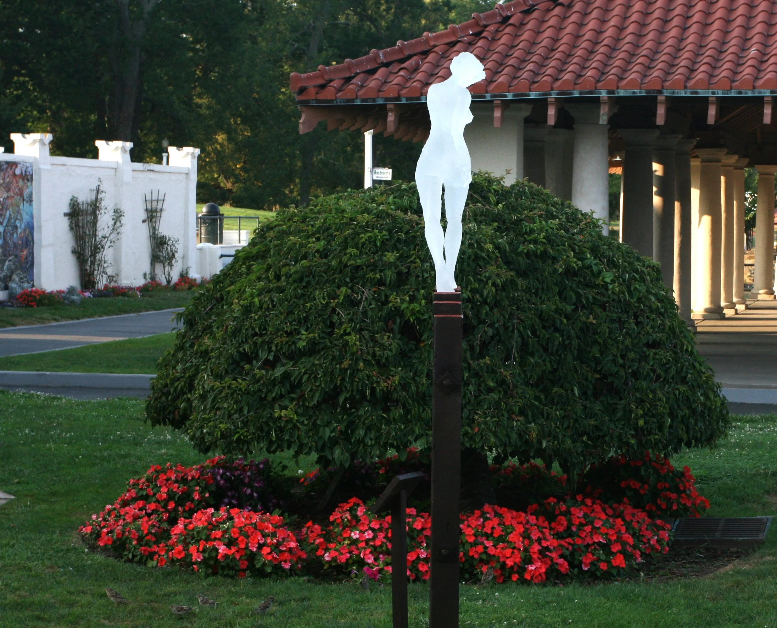 A luminous white silhouette of a sculpture of a walking woman on a tall platform next to a garden with red flowers, with a building featuring red-tiled roof and columns in the background.