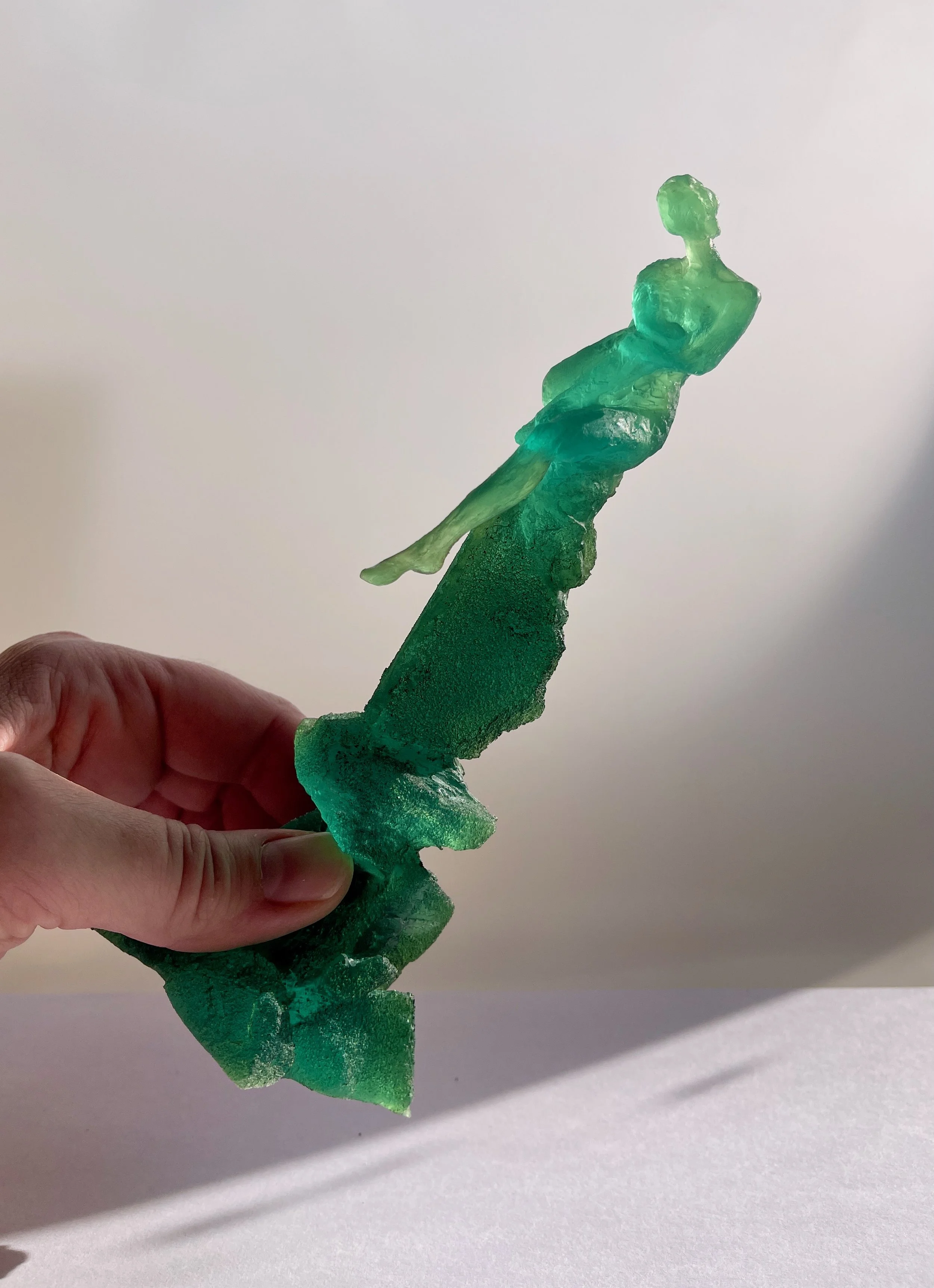 Hand holding a small green glass like sculpture of a woman sitting holding one knee up.