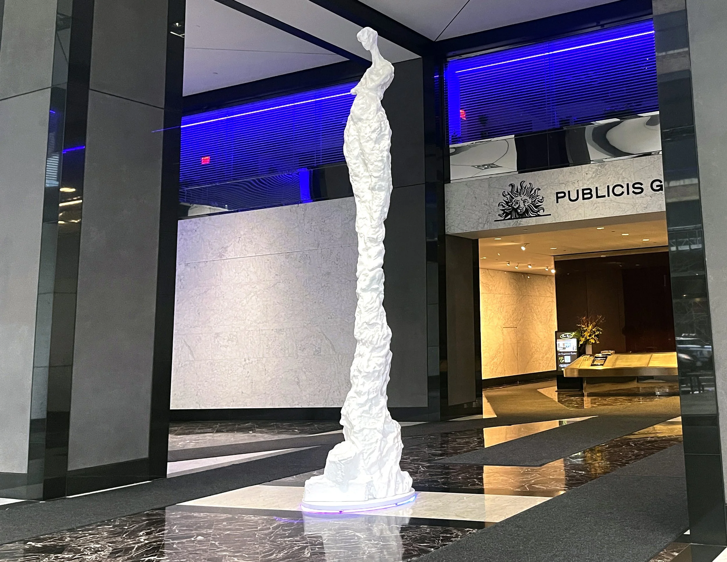 An abstract white sculpture resembling a tall, slender figure stands inside a modern building. The sculpture is illuminated from below with a purple light. The background shows part of a lobby with a marble floor, a reception desk, and a sign for Pub