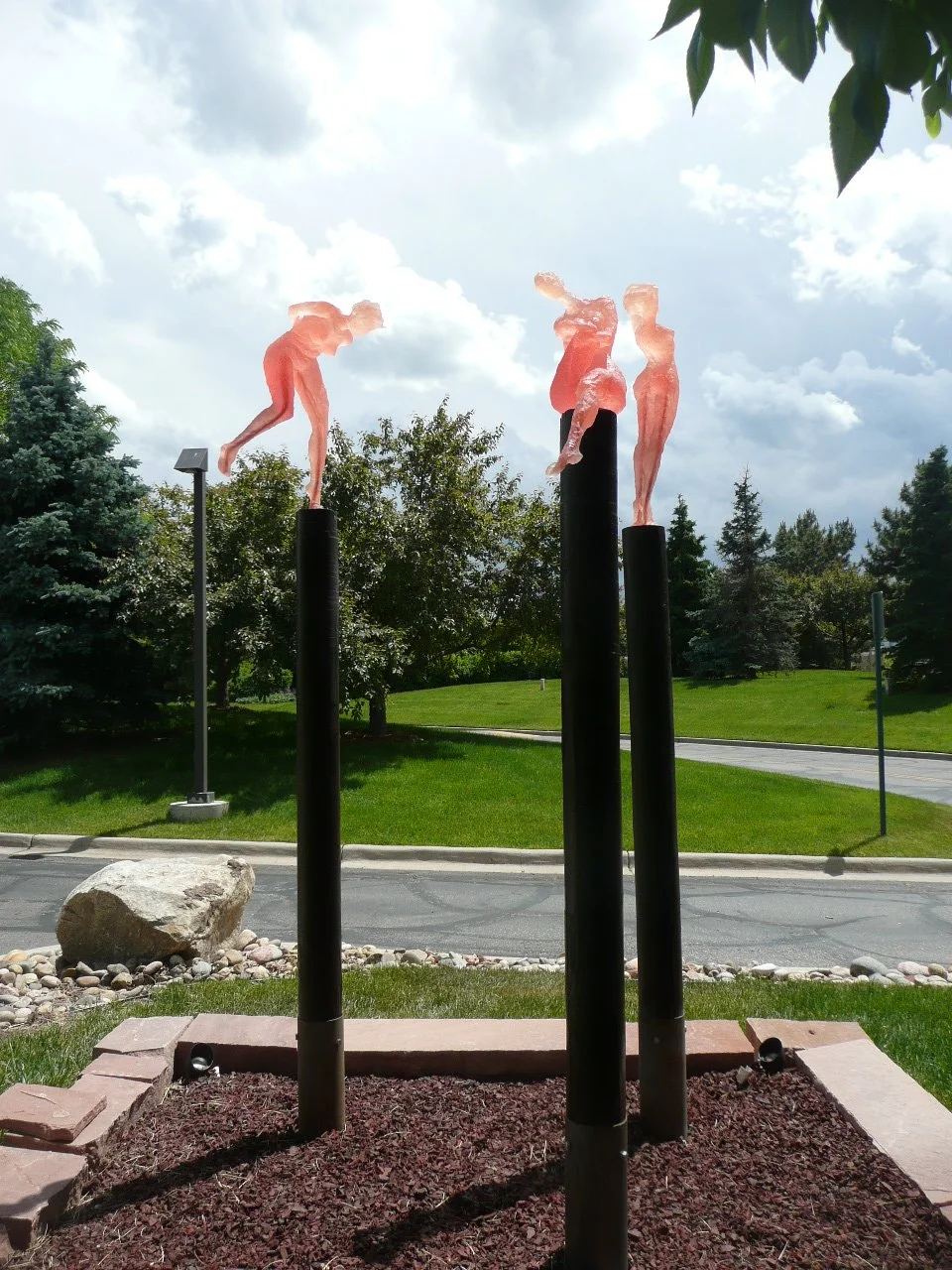 Outdoor sculpture of three abstract human figures made of translucent pink material, standing on tall black poles against a cloudy sky background.