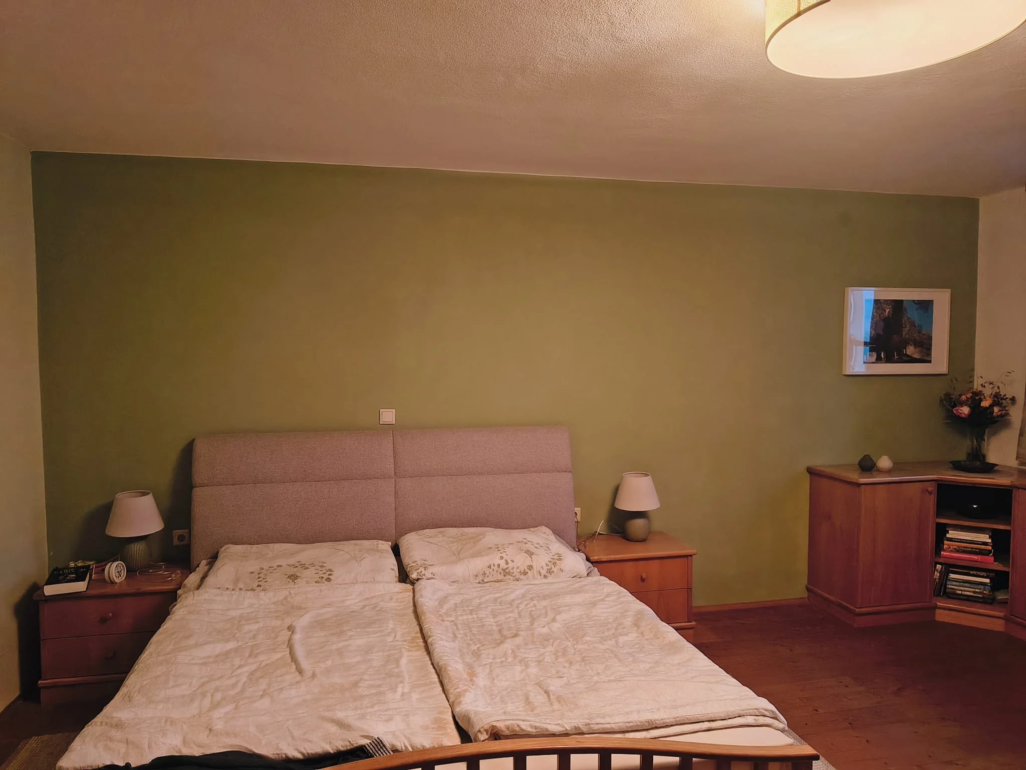 Schlafzimmer mit grünem Wand, zwei Nachttischen mit Lampen, einem Bett mit beige Decken, einem Holzschrank mit Bücher und Klammern, einem gerahmten Bild und einer Vase mit Blumen.