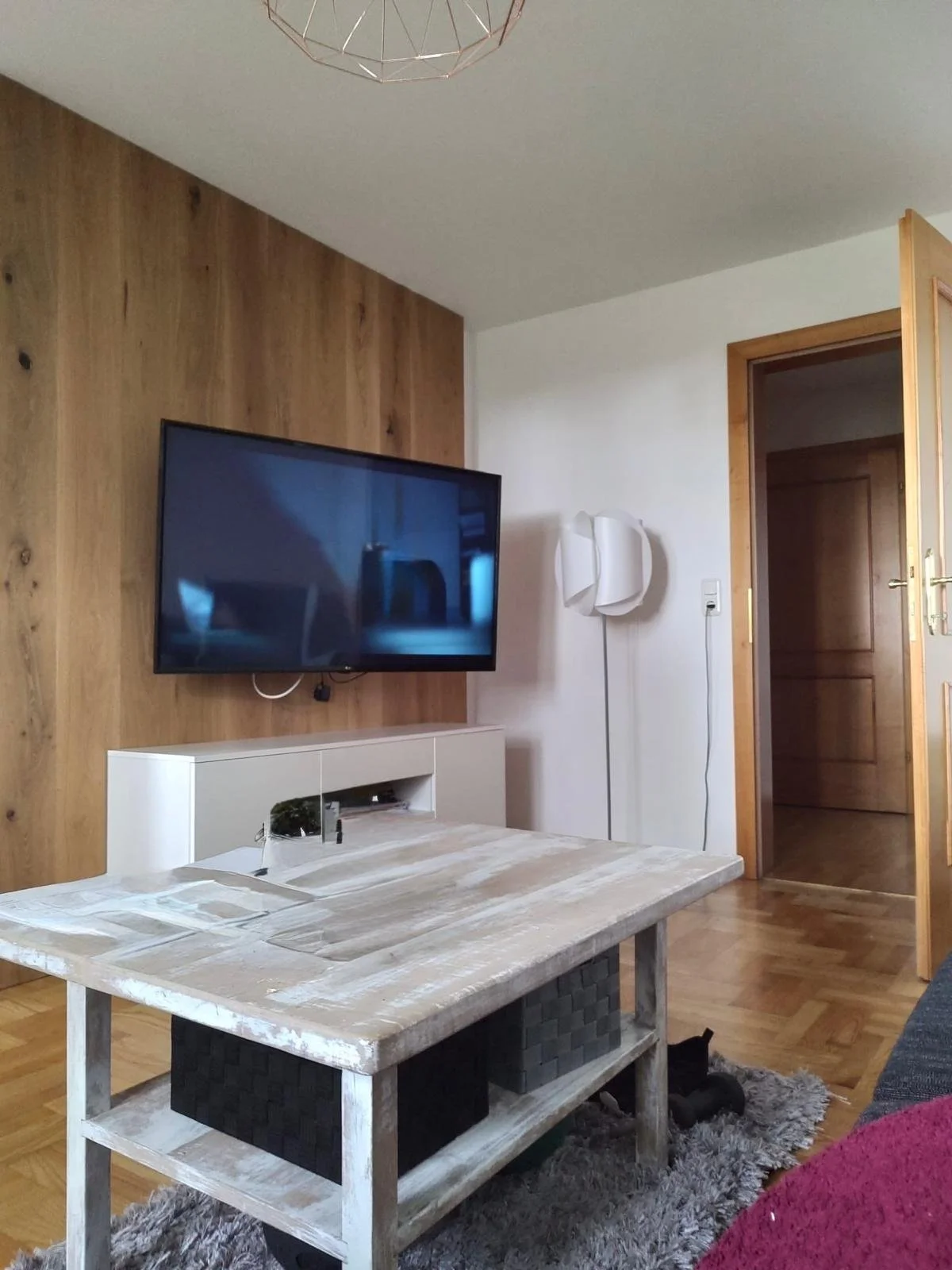 Wohnzimmer mit Holzpaneelwand, Fernseher an der Wand, weißer TV-Schrank, Stehlampe, geölter Holzfußboden, offener Holzinnentür, Teetisch, teppich, Couch mit rotem Kissen, schwarze Katze unter dem Tisch.