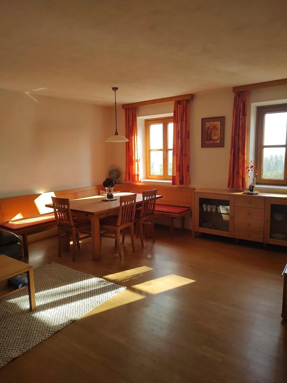 Ein hell erleuchteter Essbereich in einem Wohnzimmer, mit Holzfußboden, einem Esstisch mit vier Stühlen, bunten Vorhängen an den Fenstern und einer Glastür, die auf einen Balkon oder Garten führt.