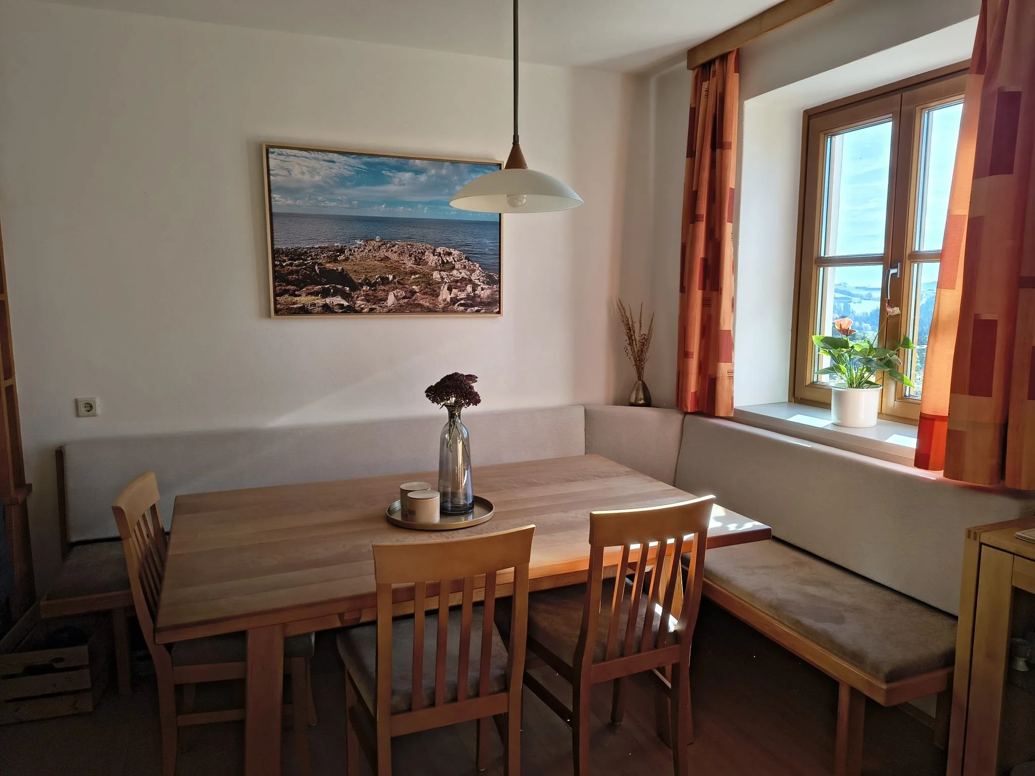 Ein heller Essbereich mit einem Holztisch, vier Holzhockern, einer Bank und einer Fensterbank mit Pflanze, helles Licht, Wandbild mit Meereslandschaft, Vorhänge in Rot-Orange-Tönen.