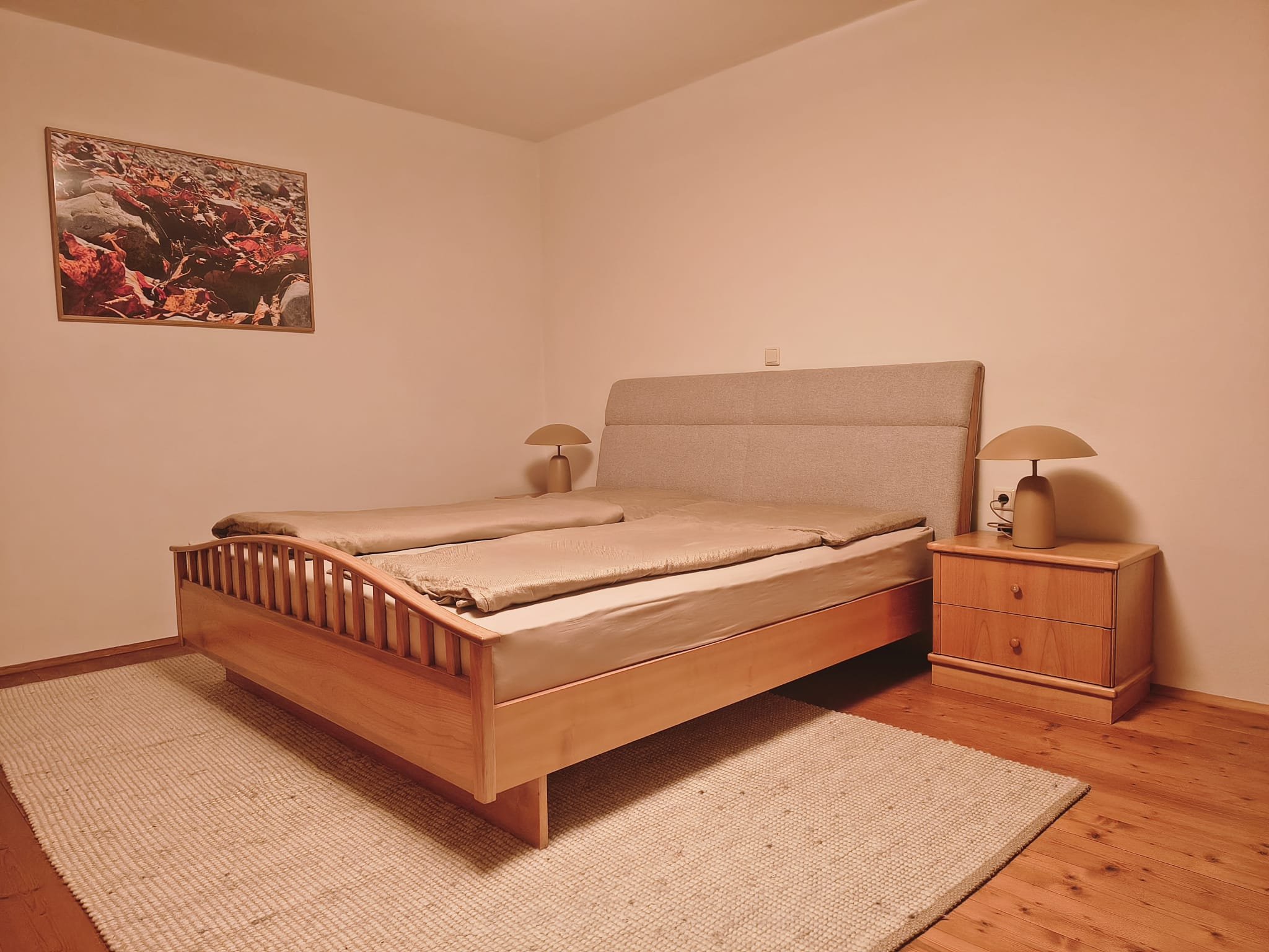 Schlafzimmer mit Doppelbett, zwei Nachttischen mit Lampen, Bild an der Wand, Holzboden und Teppich.
