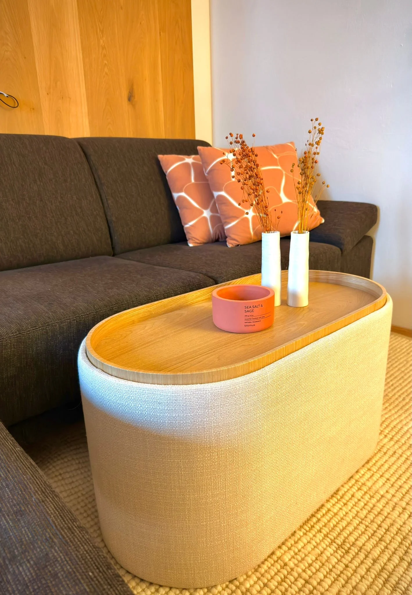 Wohnzimmer mit braunem Sofa, zwei pinken Kissen, einer weißen und einer beigen Vase mit getrockneten Zweigen, einem ovalen Couchtisch aus hellem Holz und einem weichen Teppich.
