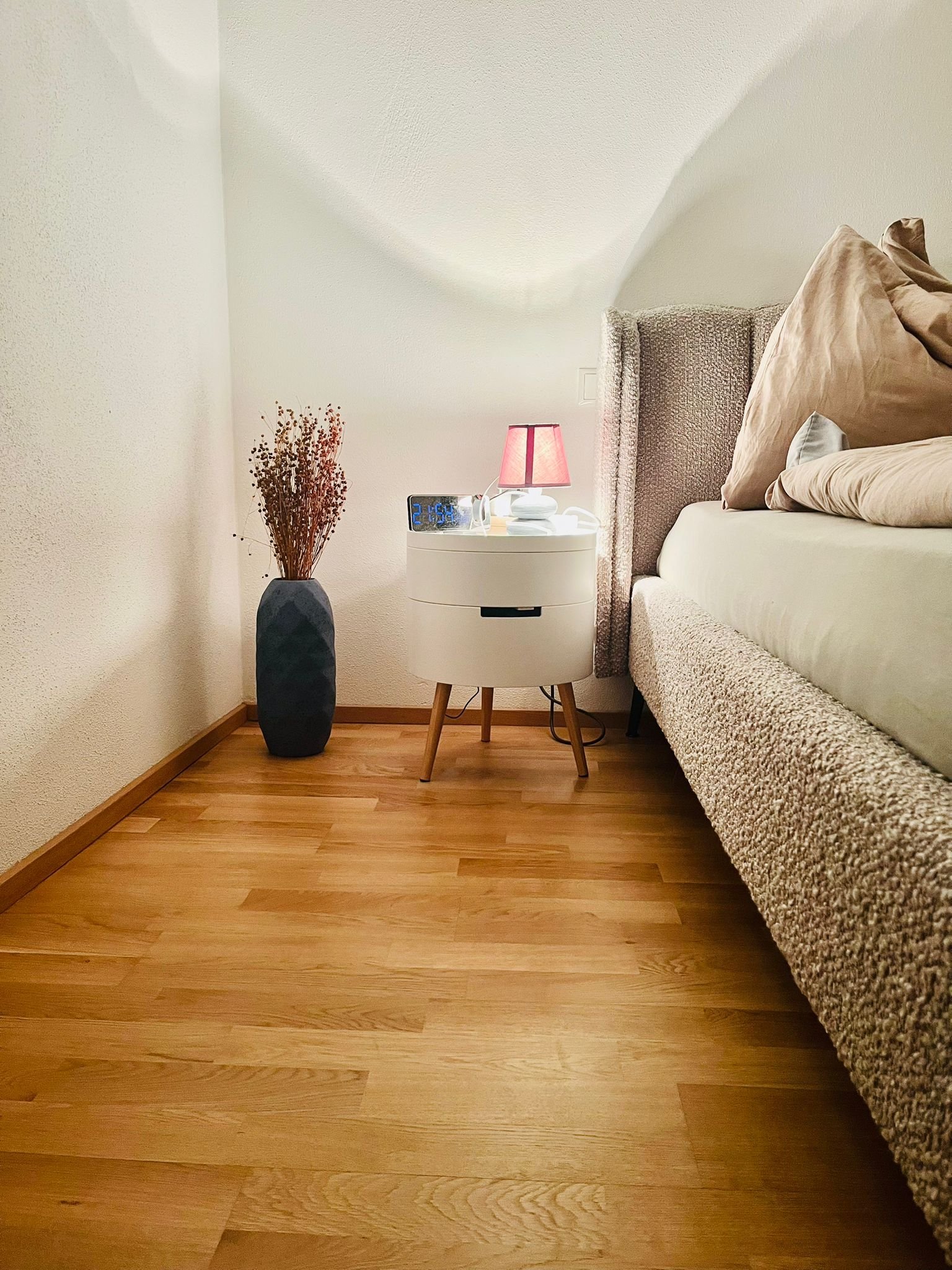 Ein Schlafzimmer mit Holzfußboden, einem weißen Nachttisch, einer Leselampe mit rotem Schirm, einem digitalen Wecker, einem symbolischen Luster, einem großen Bett mit beige- und braunfarbenen Kissen und einer dekorativen Vase mit trockenen Zweigen.