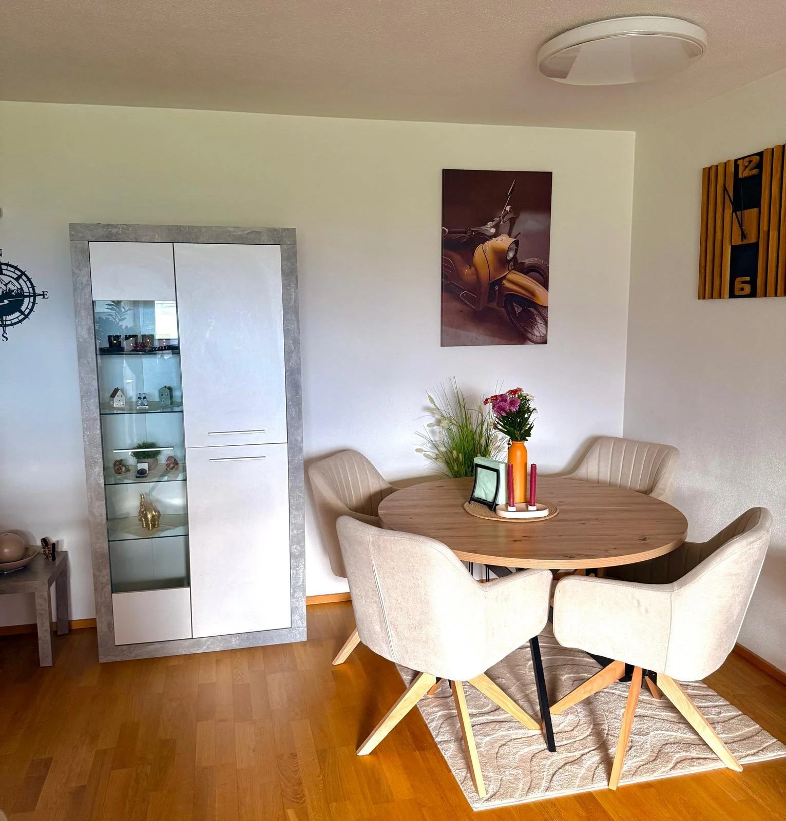 Ein esszimmer mit einem runden Holz Tisch, um den vier beige gepolsterte Stühle stehen. Auf dem Tisch steht eine Vase mit pinken Blumen und fünf Kerzen. An der Wand hängen ein Bild eines gelben Rollers und eine Uhr. Ein Weißes Möbelstück mit Glastüren steht an der Wand, daneben ein kleiner Beistelltisch. Der Boden ist aus Holz, und im Raum ist eine Deckenleuchte.