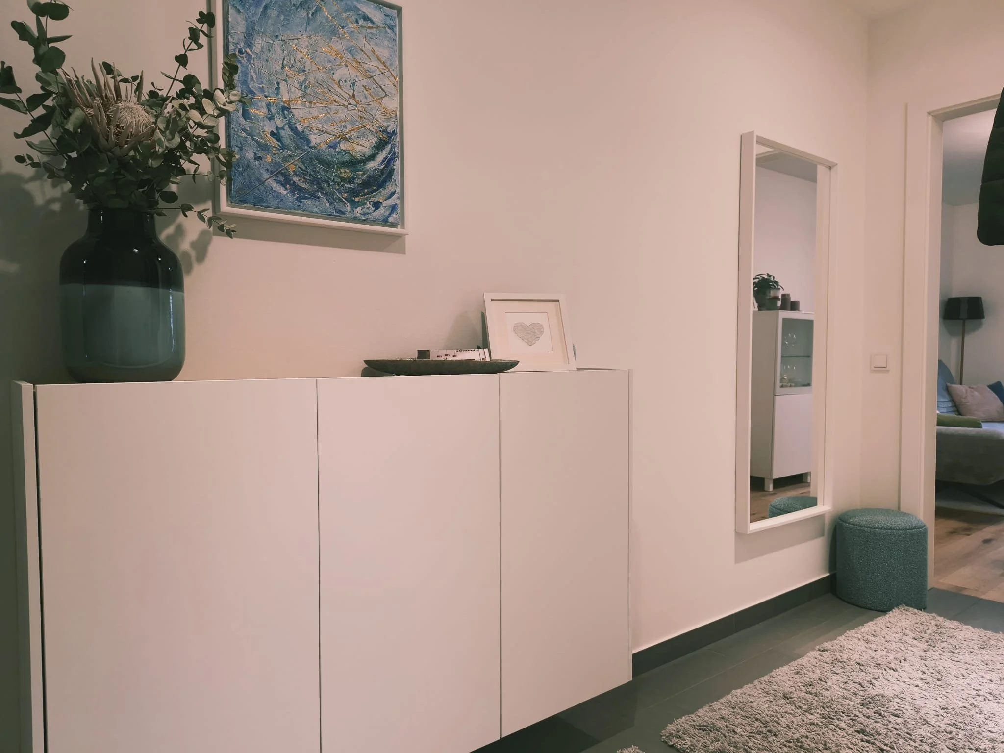 Ein weißer Sideboard mit einer Vase mit grünen Zweigen, einem Rahmen mit Herzbild und einem Ablagebrett, an der Wand hängt ein abstraktes Bild in Blau und Gold, im Hintergrund sieht man eine Türöffnung und ein Wohnzimmer mit Sofa.