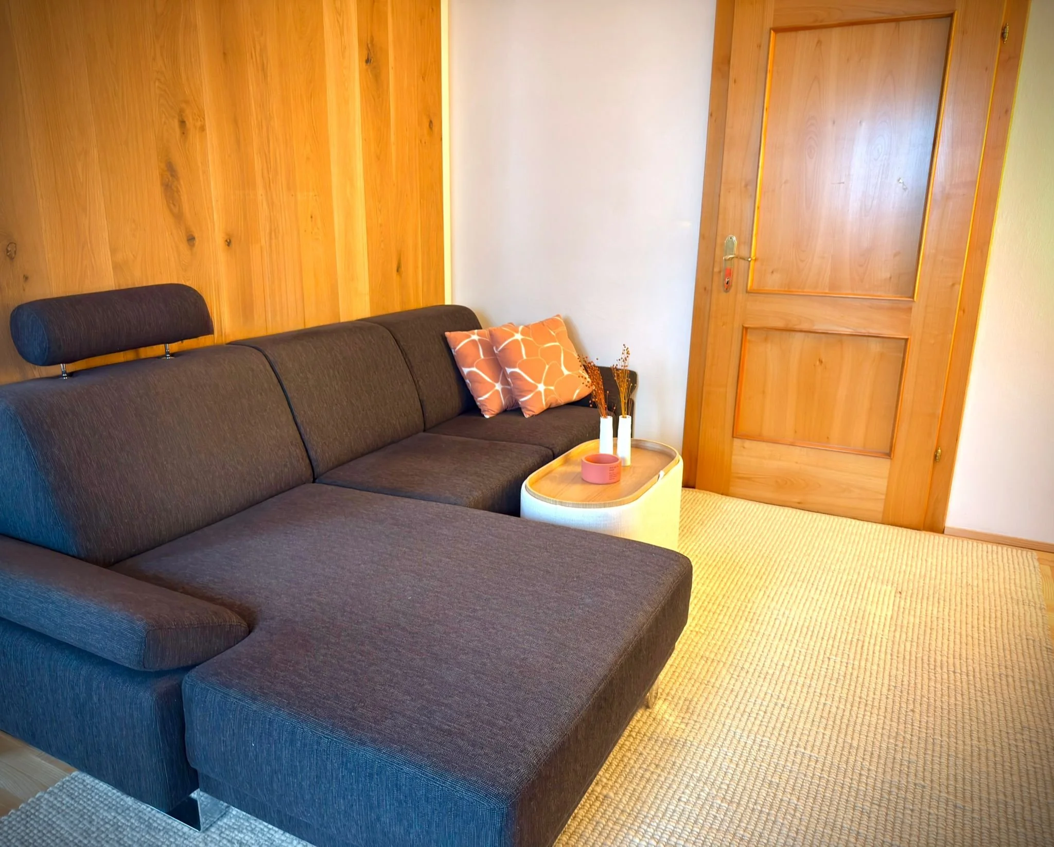 Ein Wohnzimmer mit grauem Sofa, zwei orange-weiße Kissen, einer weißen Kaffeetisch mit Vasen und Dekorationen, Holzvertäfelung an der Wand und einer Holztür.