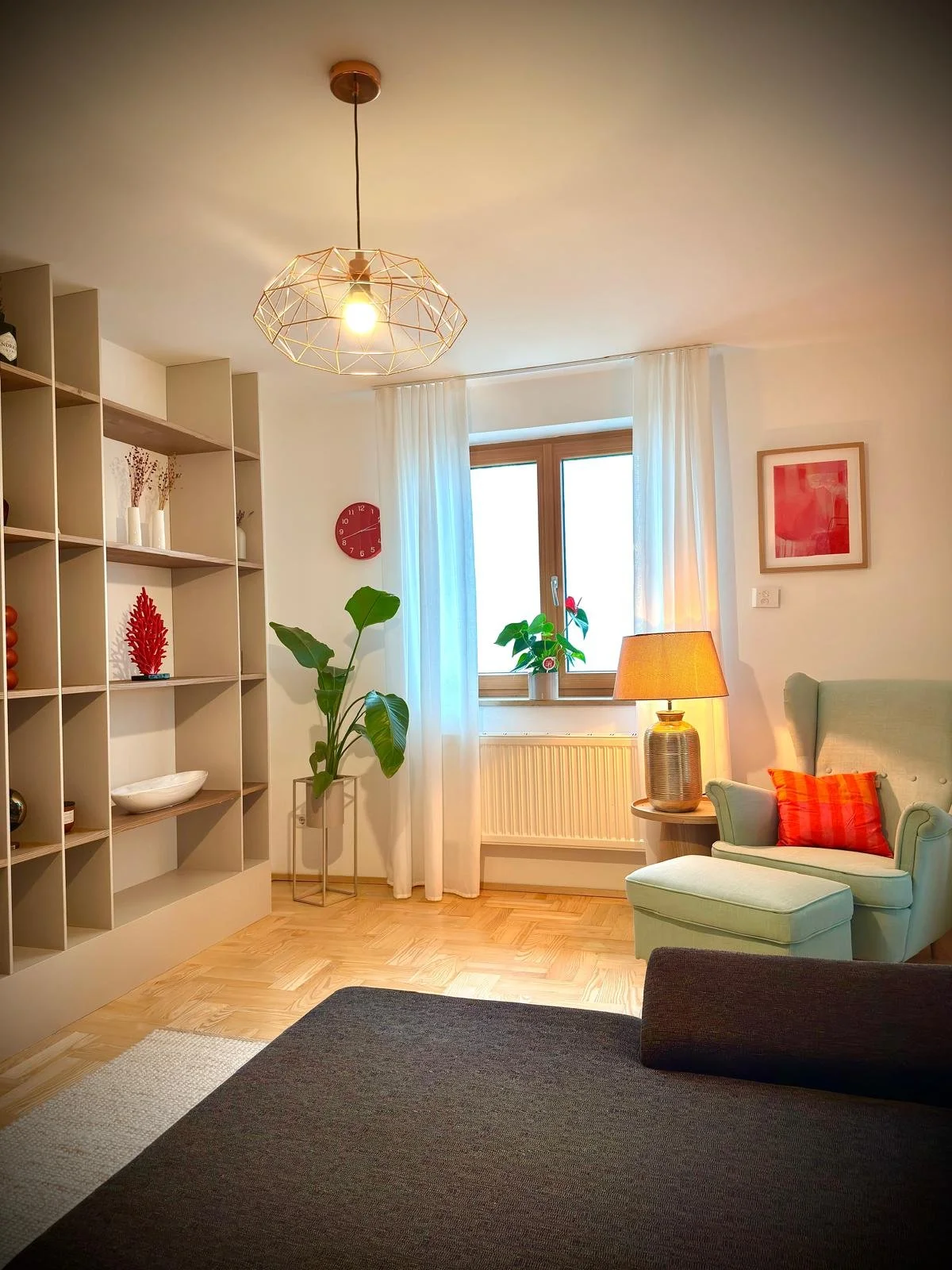 Gemütliches Wohnzimmer mit Holzfußboden, hellgrüner Sessel mit roter Kissen, Stehlampe, Fenster mit weißen Vorhängen, Pflanzen, dekorativen Gegenständen auf einem Regal, Wanduhr und Wandbild.