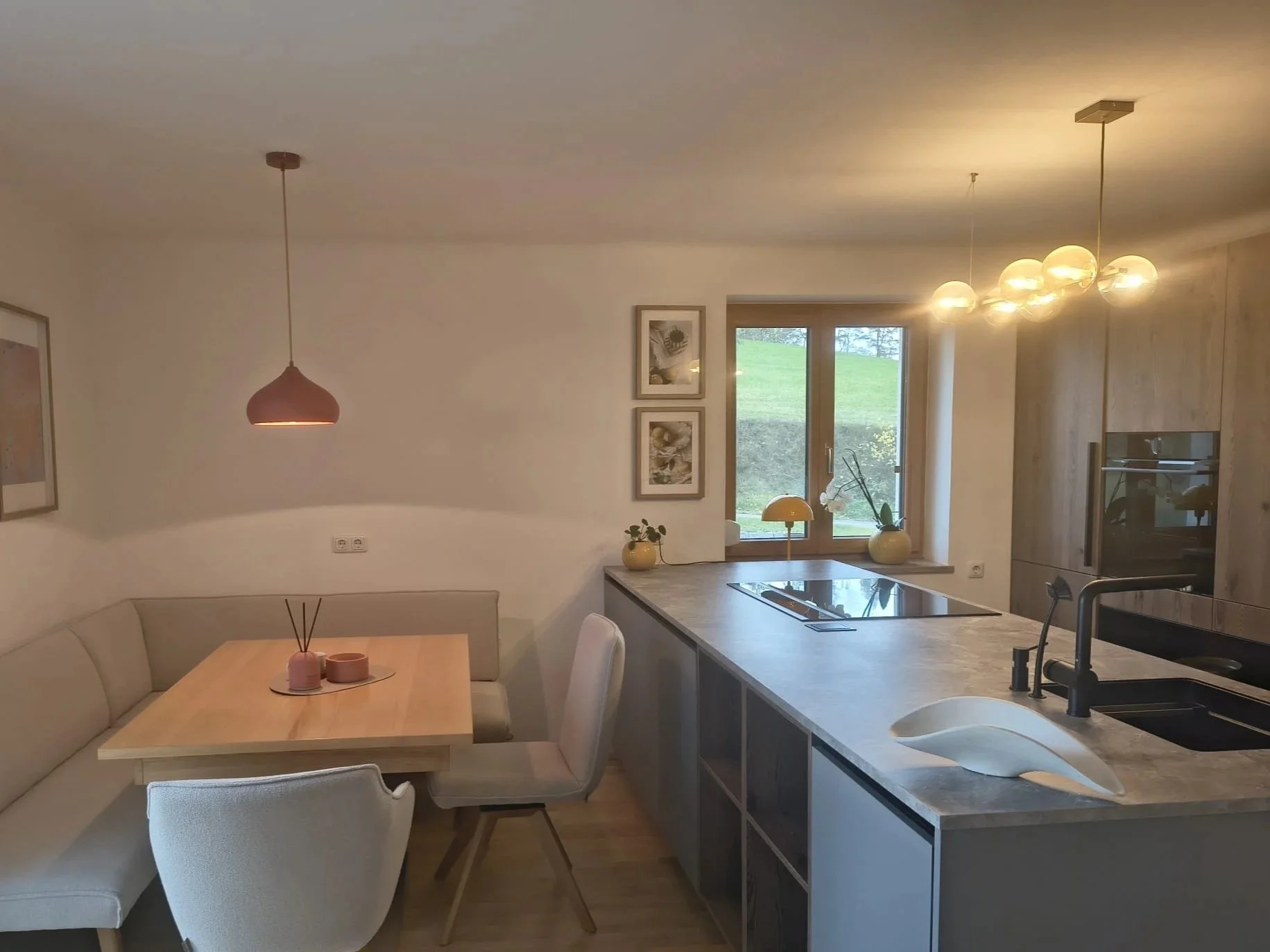 Moderne offene Küche mit elektrischem Kochfeld, Spüle, Essbereich mit Sitzbank und Stühlen, kalte Farbpalette, Pflanzen und Pendelleuchten, Fenster mit Blick ins Grüne.