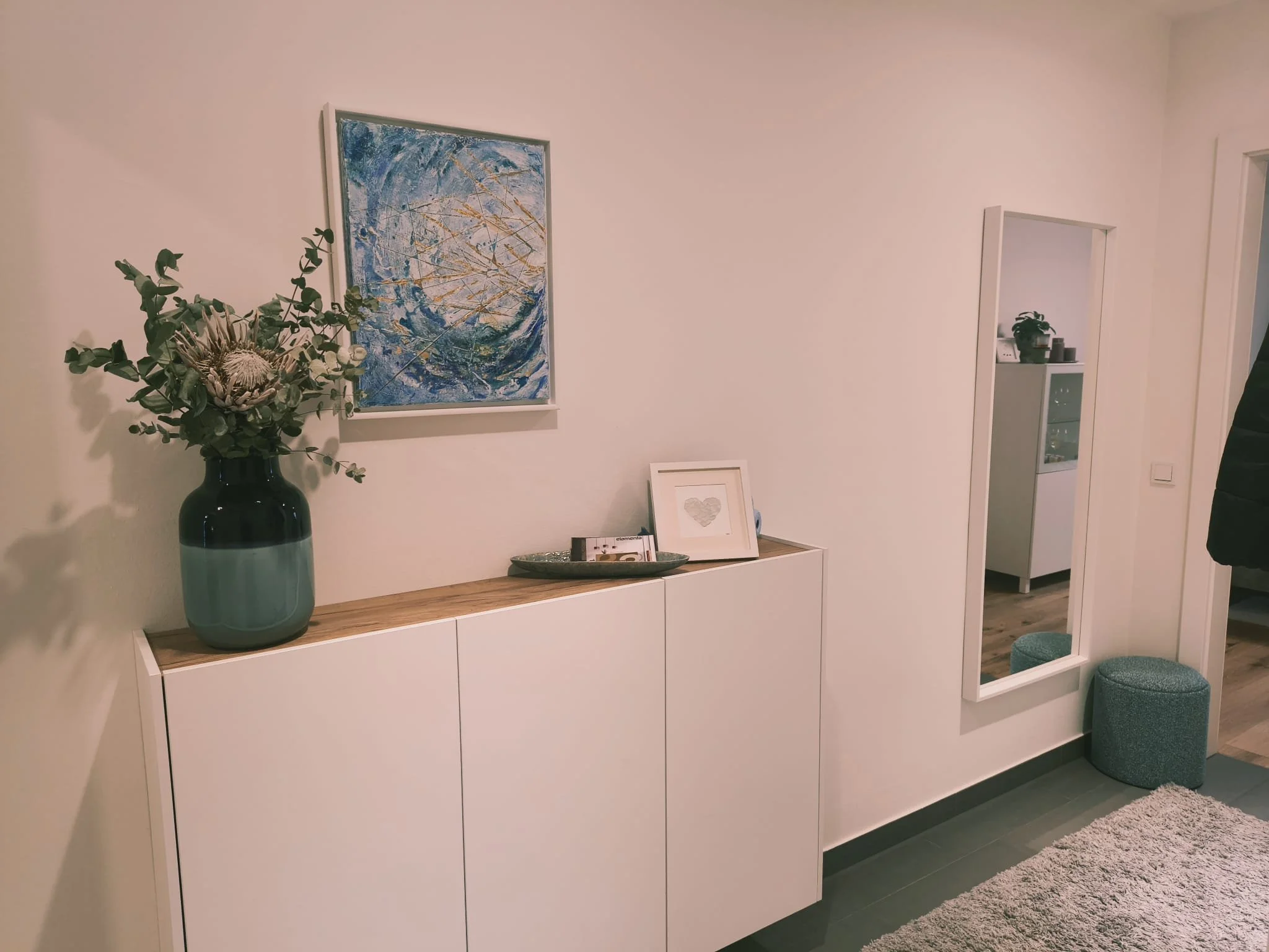 Ein Flur mit weißer Wand, einem weißen Sideboard mit Holztop, einer Spitze einer großen Vase mit grünen Pflanzen, einem gemalten Bild und einer kleinen Bilderrahmen- und Deko-Schale auf dem Sideboard. Es gibt einen Spiegel an der Wand und eine kleine gepolsterte Ottomane. Im Hintergrund sieht man einen Raum mit dunklem Boden, einer weißen Schrankwand und Pflanzen.