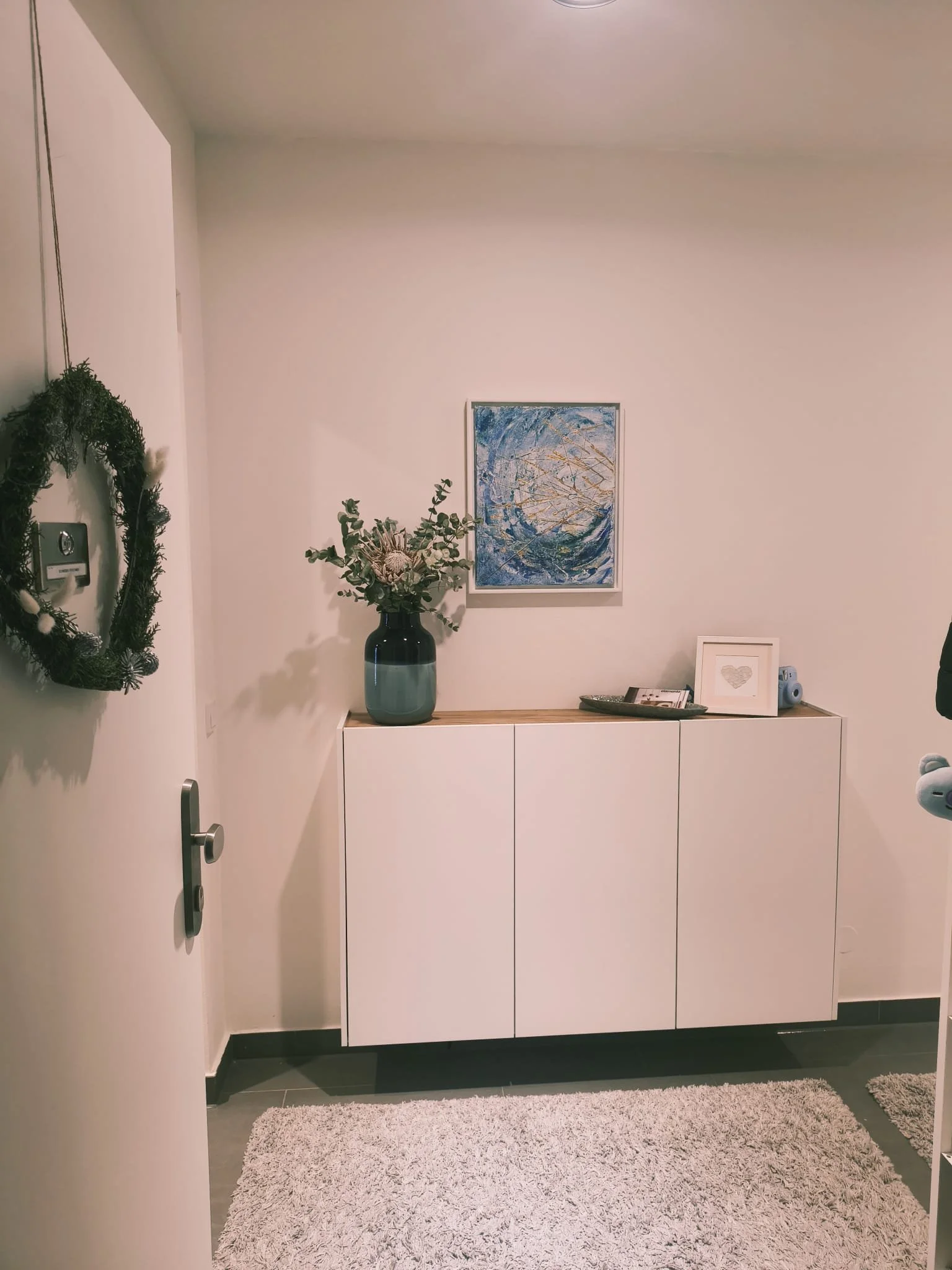 Ein Flur mit weißem Chemiefaser-Gegenstand, einer blauen Vase mit Blumen, einem Bild, und einem weißen Schrank mit Holzplatte.