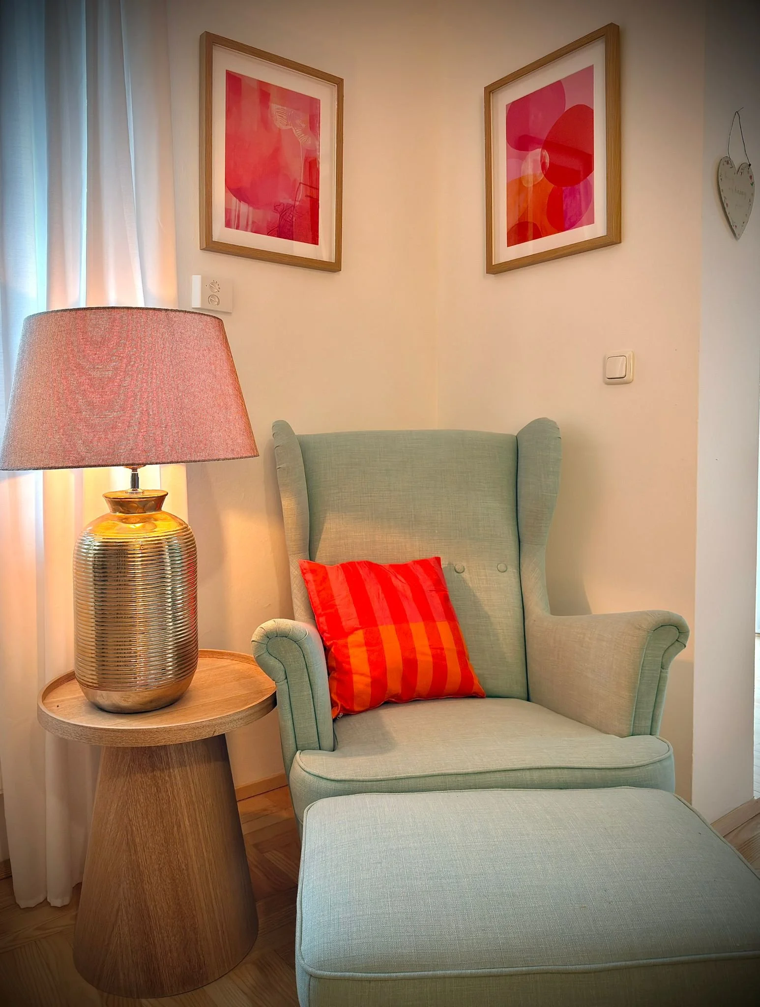 Gemütliches Wohnzimmer mit einem beigen Sessel, einer roten Kissen und einem Beistelltisch mit einer goldenen Lampe, Wandbilder mit rotem und pinkem Kunstwerk, und weißen Vorhängen.