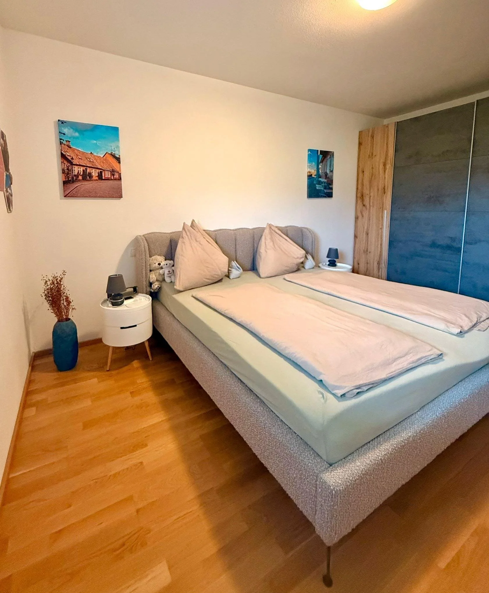 Ein Schlafzimmer mit einem großen, gepolsterten Bett, zwei Nachttischen, Wandbilder, Holzboden und einer großen Einbauschrankwand.