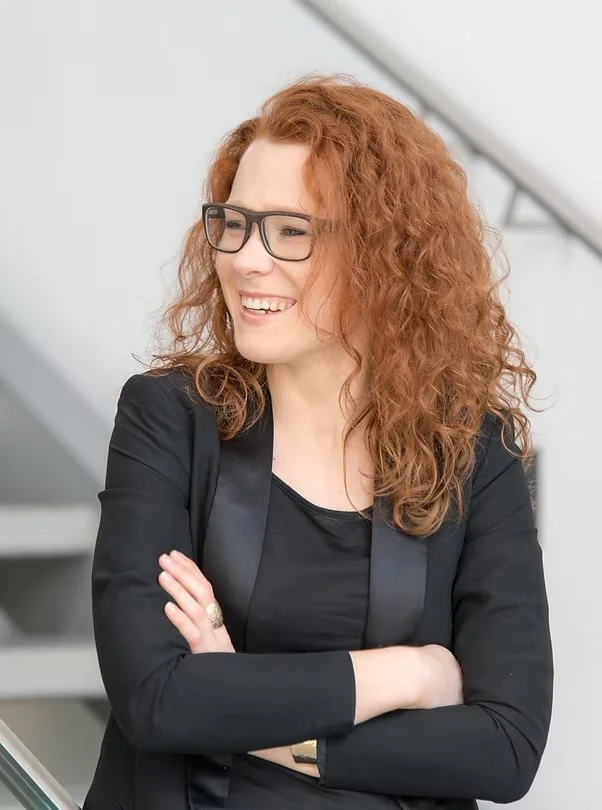 Eine lächelnde Frau mit lockigem roten Haar, Brille und schwarzer Jacke steht mit verschränkten Armen vor einer weißen Wand im Innenraum.
