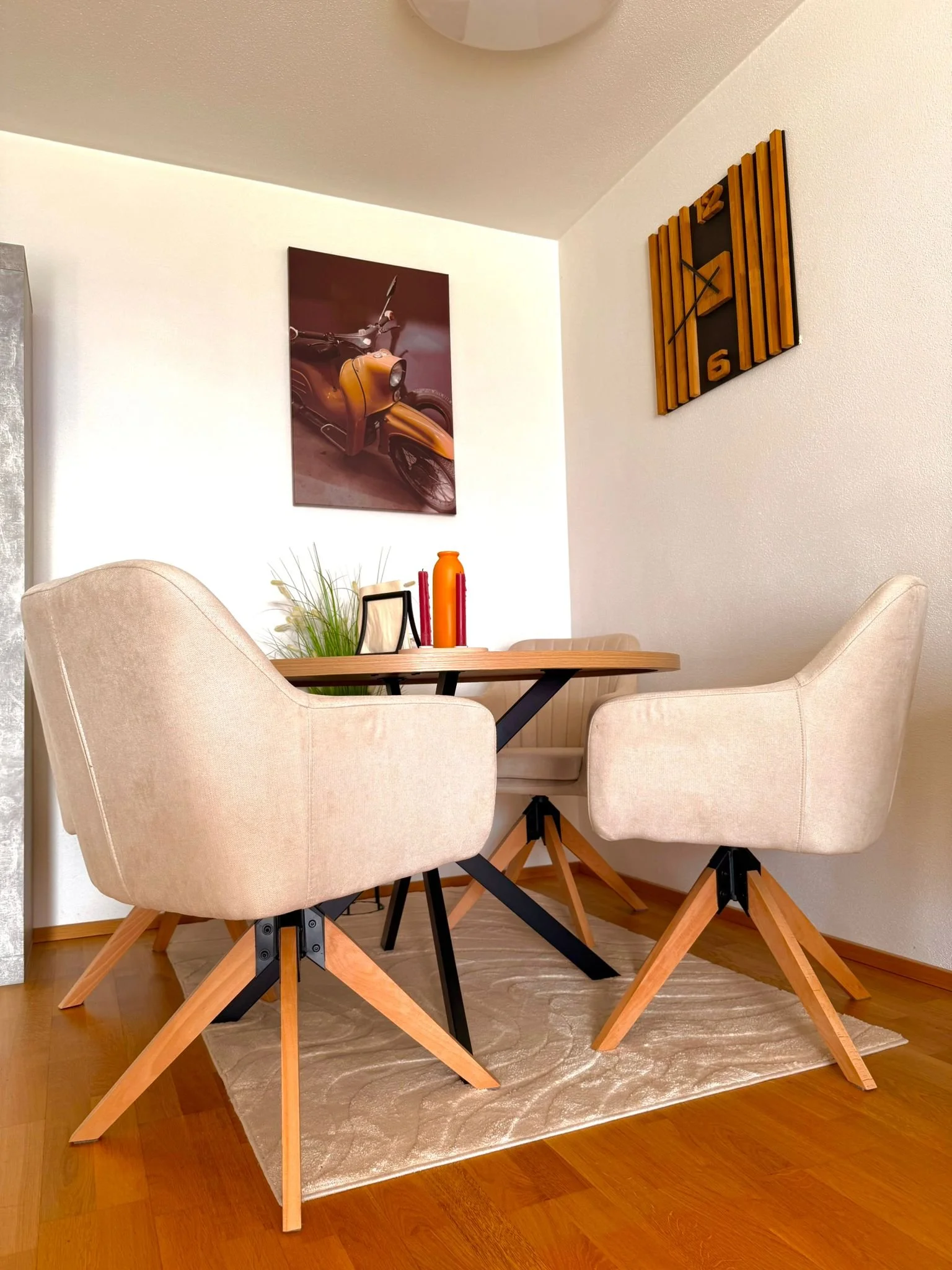 Moderner Essbereich mit einem runden Holztisch, vier beige gepolsterte Stühle mit Holzbeinen, eine Wand mit Kunstwerk eines Motorrads und eine große Uhr im Retro-Stil.