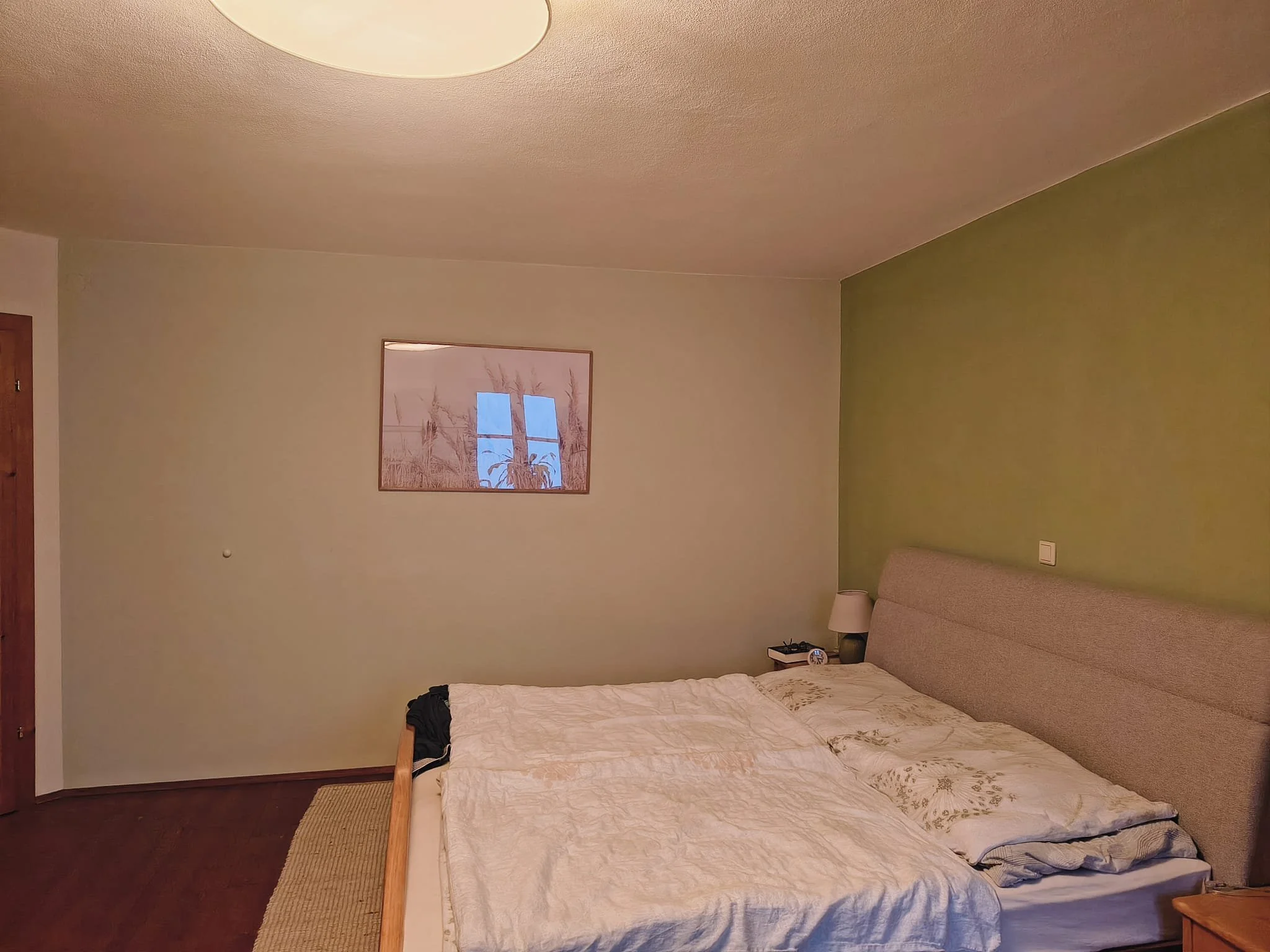Ein Schlafzimmer mit einem großen Bett, einem Nachttisch mit Lampe, einem kleinen Wecker und einem Telefon. Die Wand hinter dem Bett ist grün, die andere Wand ist beige, und es gibt ein Bild an der Wand.