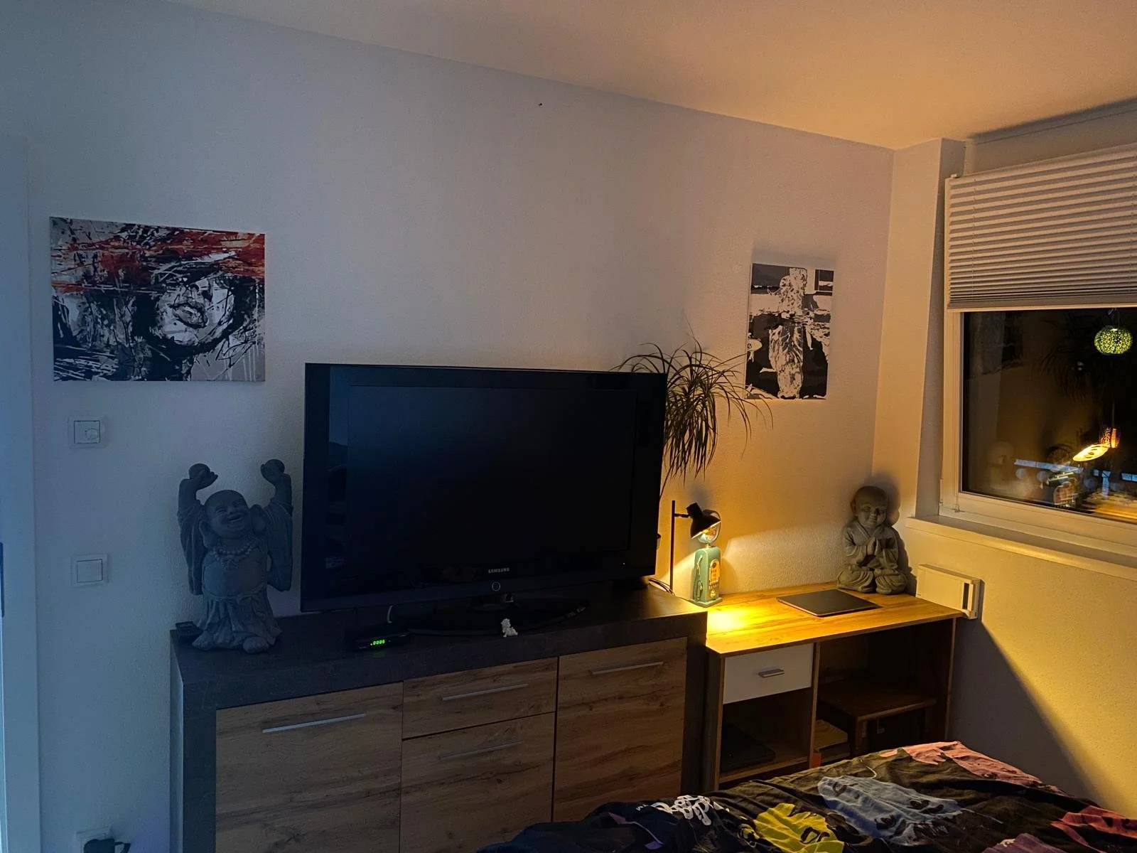 Wohnzimmer mit Fernseher, Pflanzen, Betthaupt und dekorativen Figuren auf Komoden, Wandbilder und ein Fenster