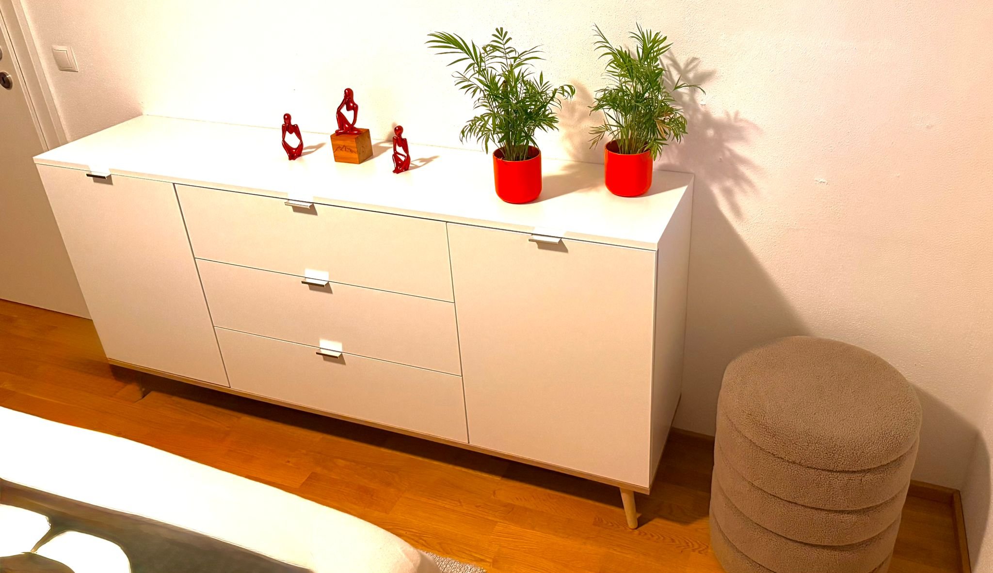 Weißer Sideboard mit drei Schubladen und zwei Türen, auf dem drei rote Deko-Figuren, zwei Topfpflanzen in roten Töpfen und eine beige runde Sitztruhe vor weißer Wand und Holzboden.