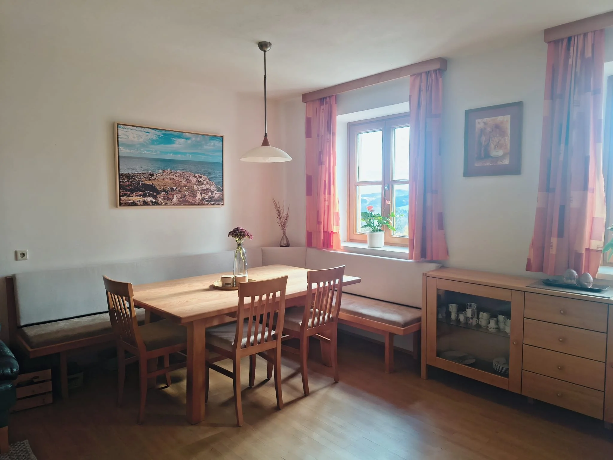 helles Esszimmer mit Holzstühlen, einem rechteckigen Tisch, einem Fenster mit roten Vorhängen, Pflanzen und Bildern an der Wand.