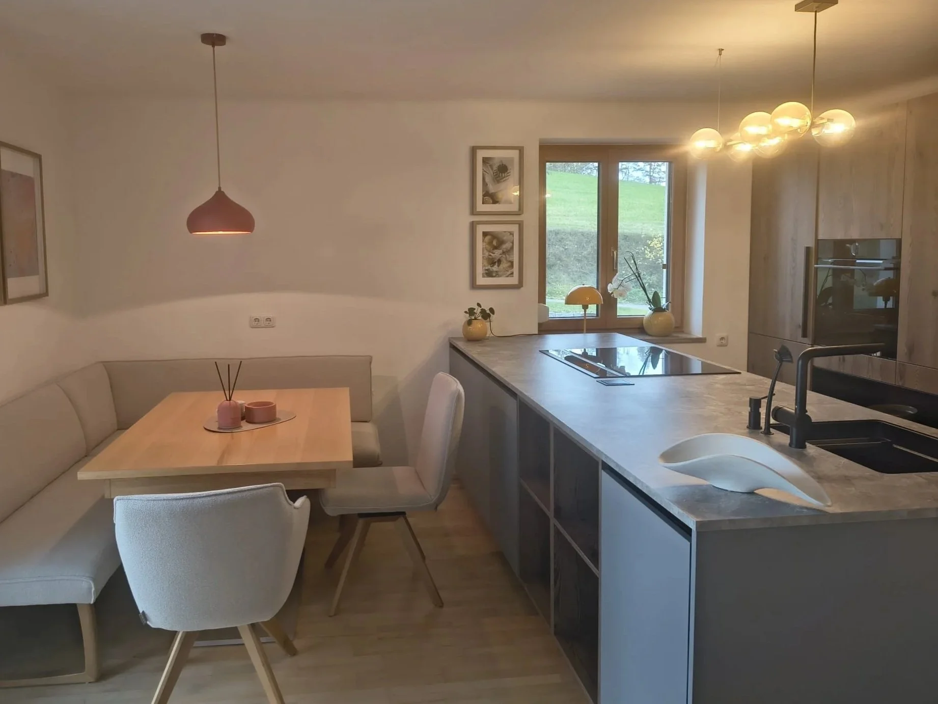 Offene Küche und Essbereich, helle Möbel, zwei Vasen mit Pflanzen auf der Fensterbank, modern gestaltete Beleuchtung