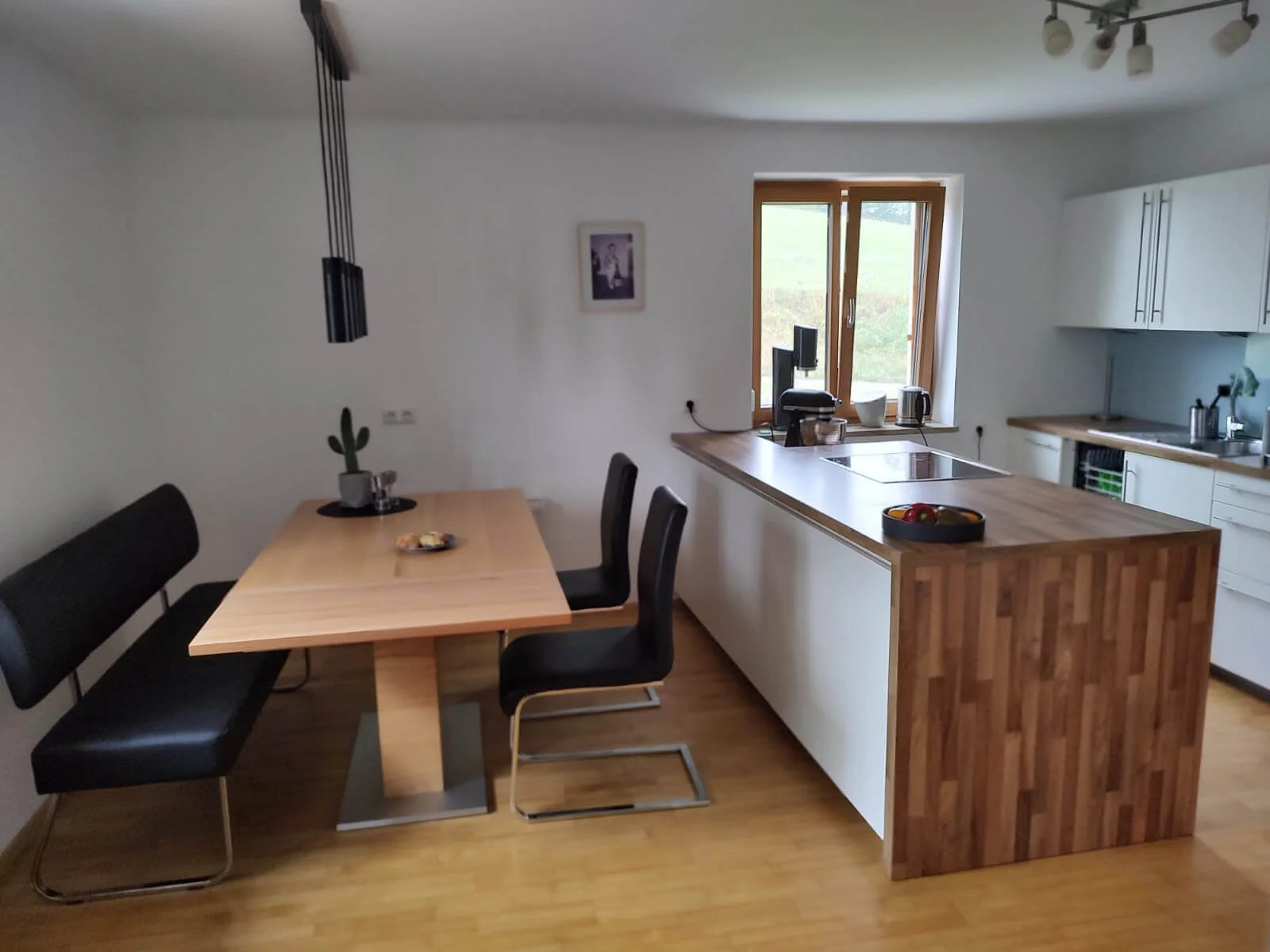 Moderner Küchen- und Essbereich mit Holztisch, schwarzen Stühlen, Küchengebäuden, einer Küchenarbeitsinsel mit Holzoberfläche, Fenster, Kaffeemaschine, Wasserkocher und Obstschale.