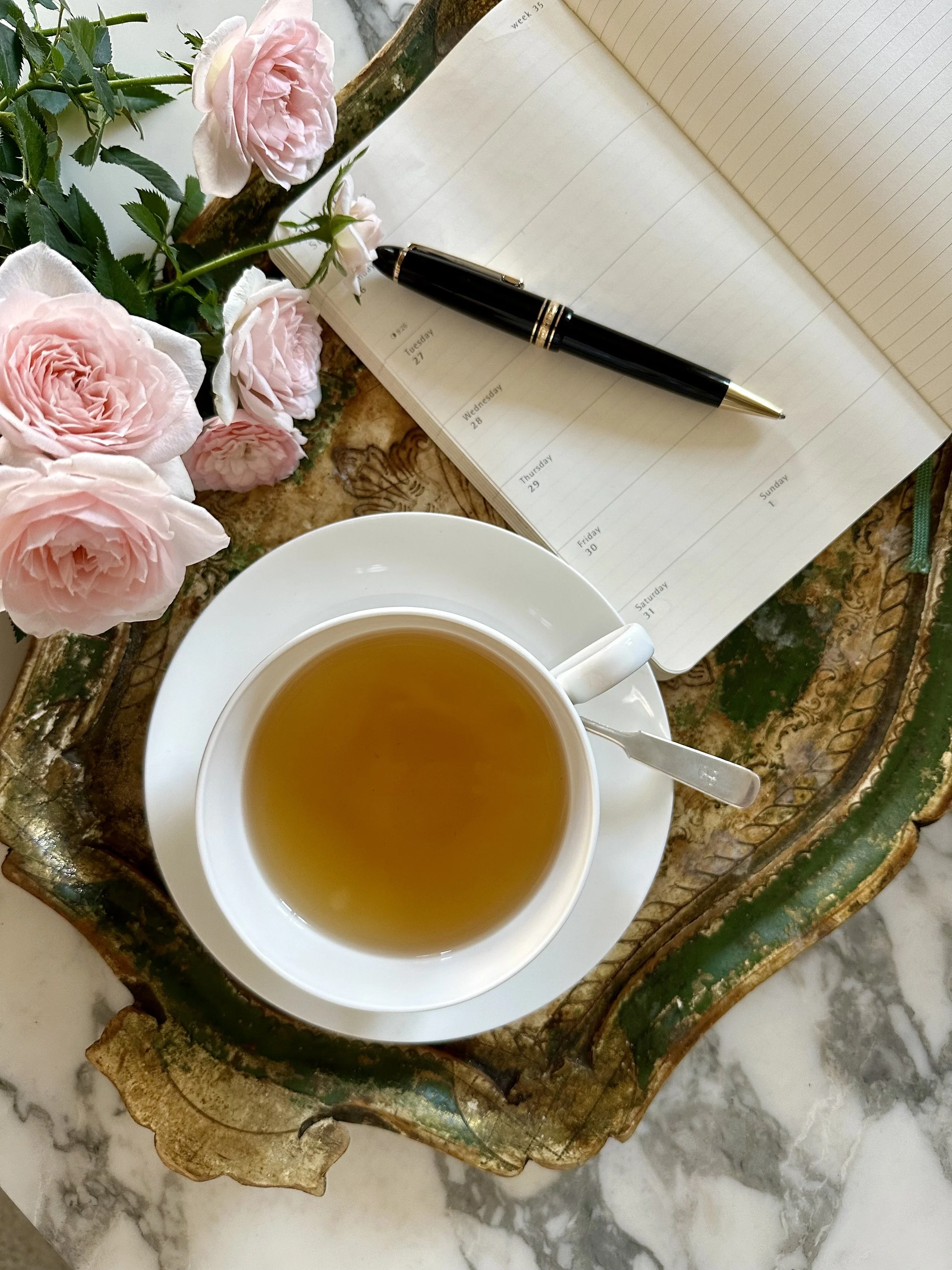 A cup of tea on a saucer with a teaspoon, an open planner with a black pen on top, pink roses, and a decorative wooden tray on a marble surface.