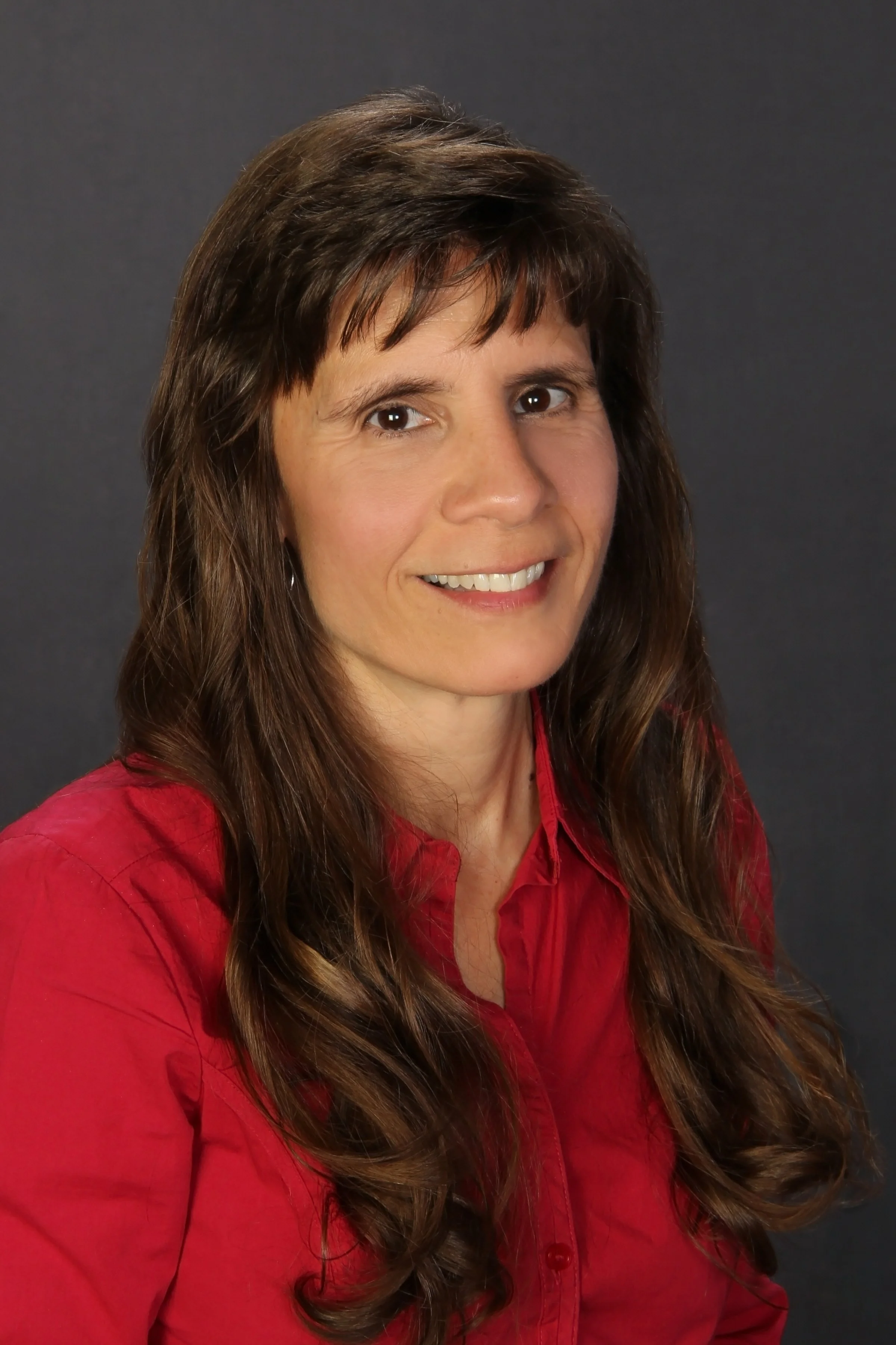 A woman with long brown hair wearing a red shirt, smiling at the camera against a dark background.
