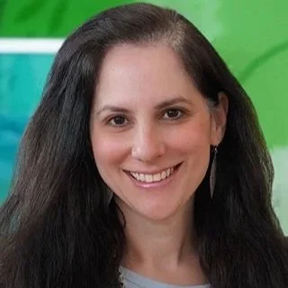 A woman with long dark hair smiling, wearing earrings, with a green background.