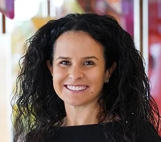 Close-up of a woman with curly black hair smiling, indoors with blurred colorful background.