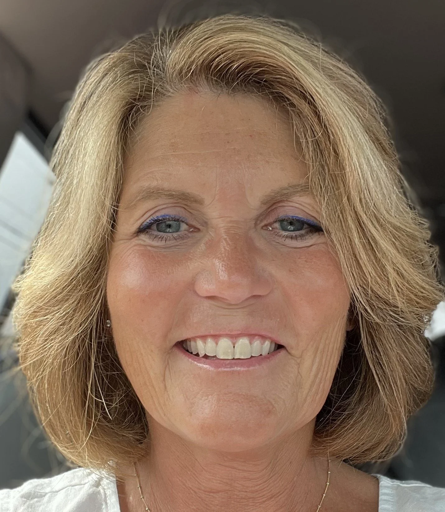 Close-up of a smiling middle-aged woman with blonde hair and blue eyes, wearing makeup, earrings, and a white top.