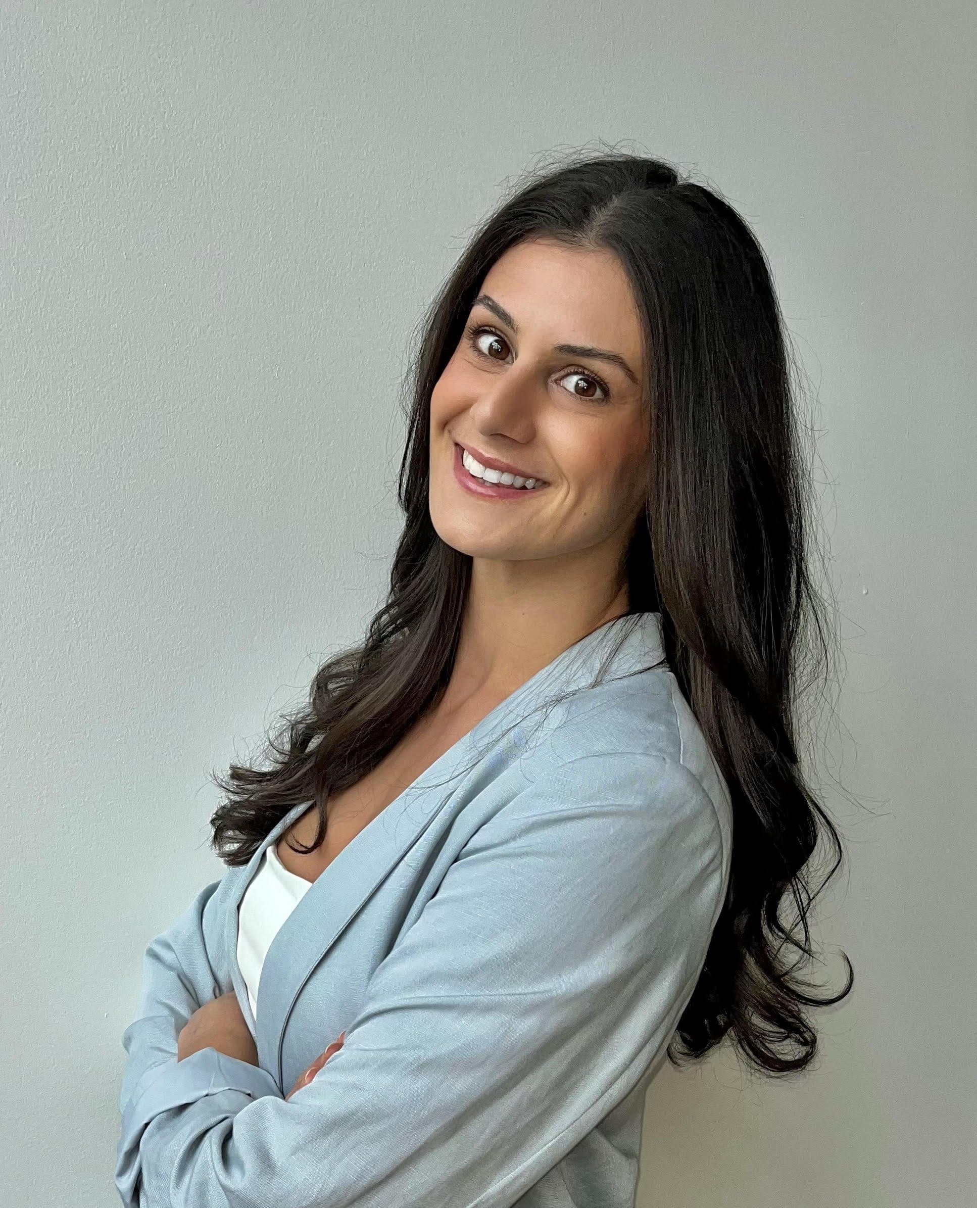 Portrait of a smiling woman with long dark hair, wearing a light gray blazer over a white top, standing against a plain beige wall.