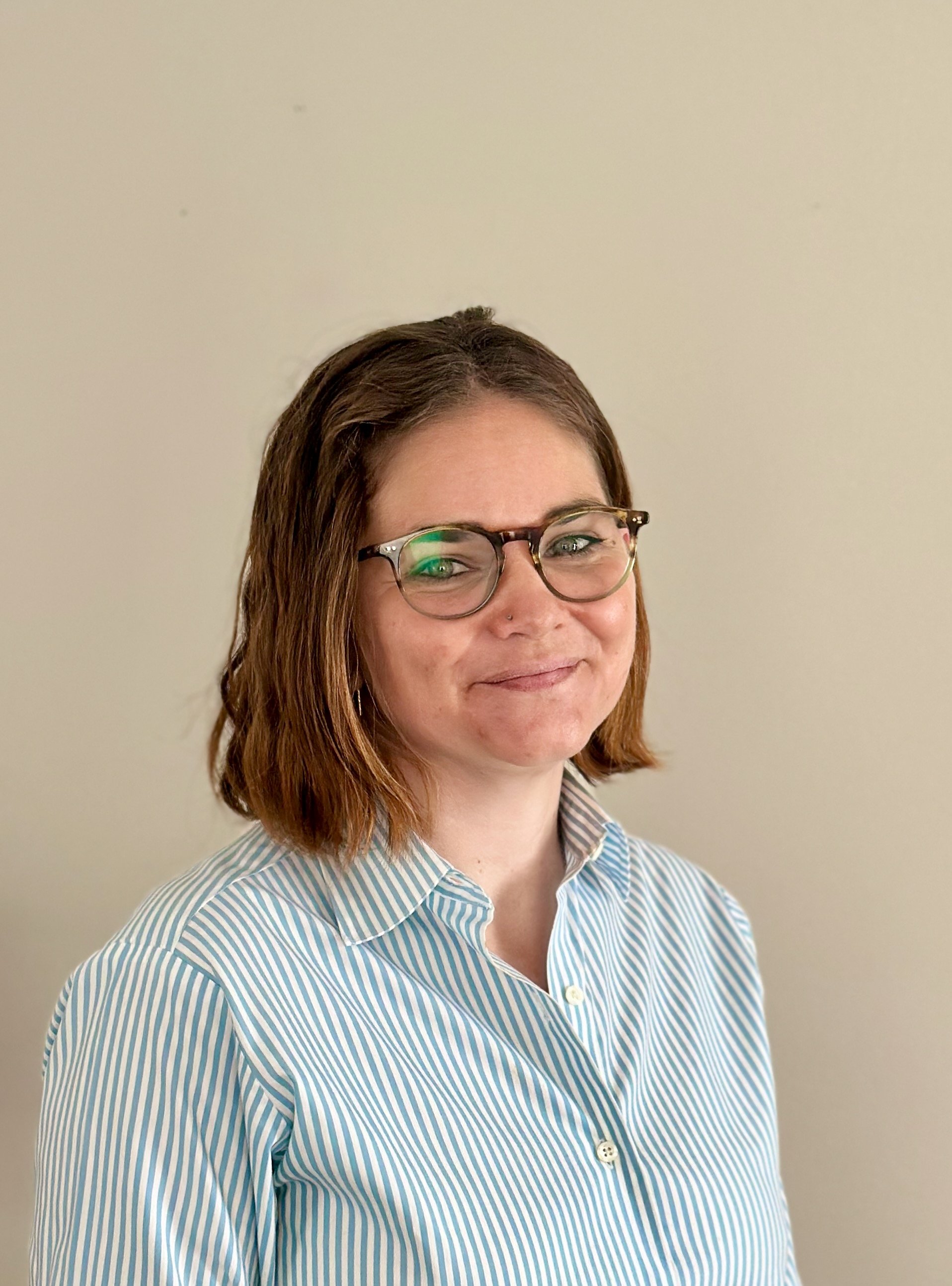 A woman with short brown hair, wearing glasses and a blue and white striped button-up shirt, smiling slightly, standing against a plain light-colored wall.