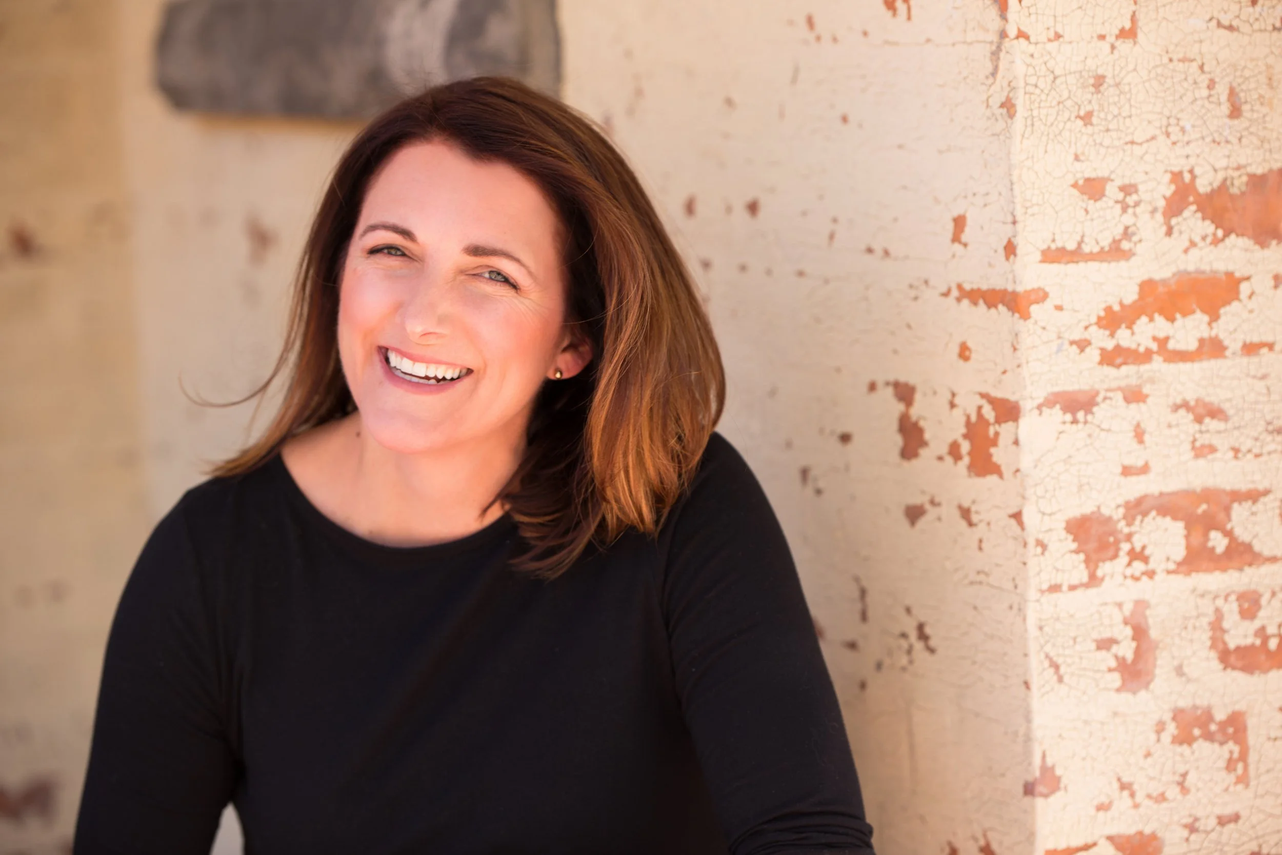 A woman with shoulder-length brown hair smiling and wearing a black top, sitting against a textured brick wall with peeling paint.