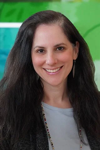 A woman with long dark hair smiling, wearing a gray top, earrings, and a necklace, with a green background.
