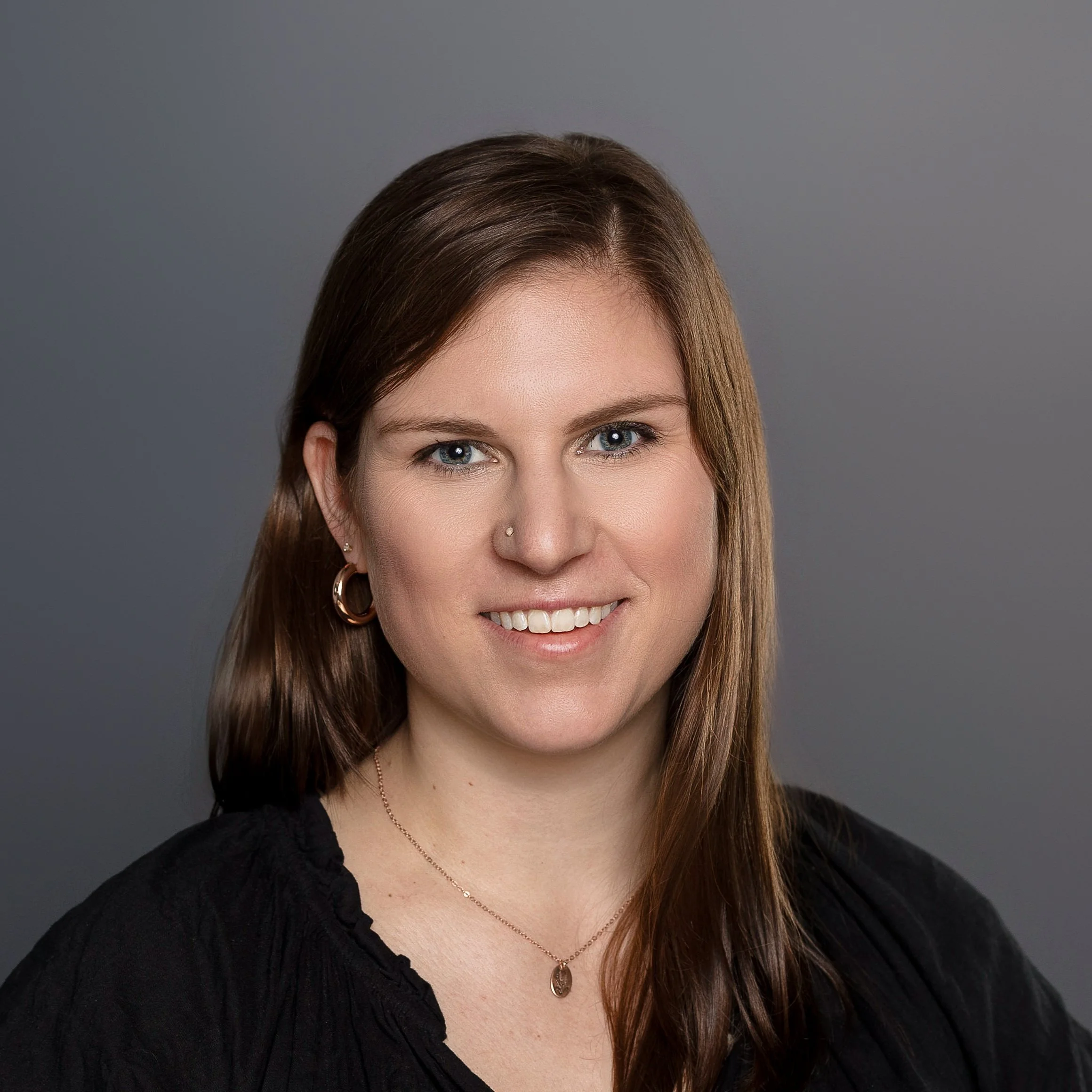Professional headshot of a woman with blue eyes and long brown hair, wearing a black top, gold hoop earrings, a gold necklace with a small pendant, and a nose piercing, smiling against a gray background.
