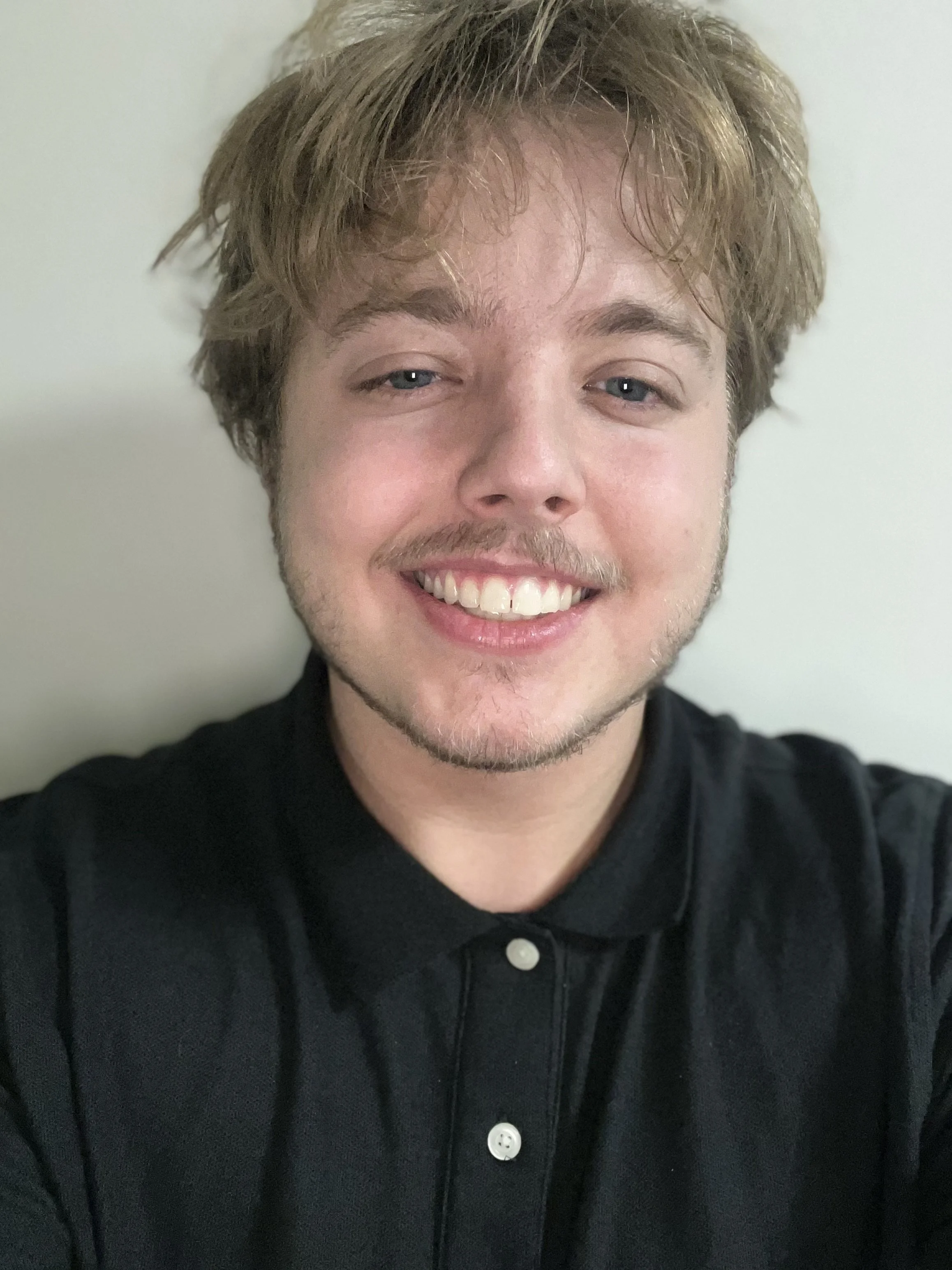 A young man with curly, light brown hair smiling and wearing a black collared shirt.