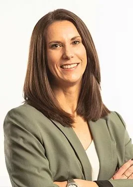 A woman with shoulder-length brown hair, smiling, wearing a green blazer over a white top, standing with arms crossed against a plain light background.