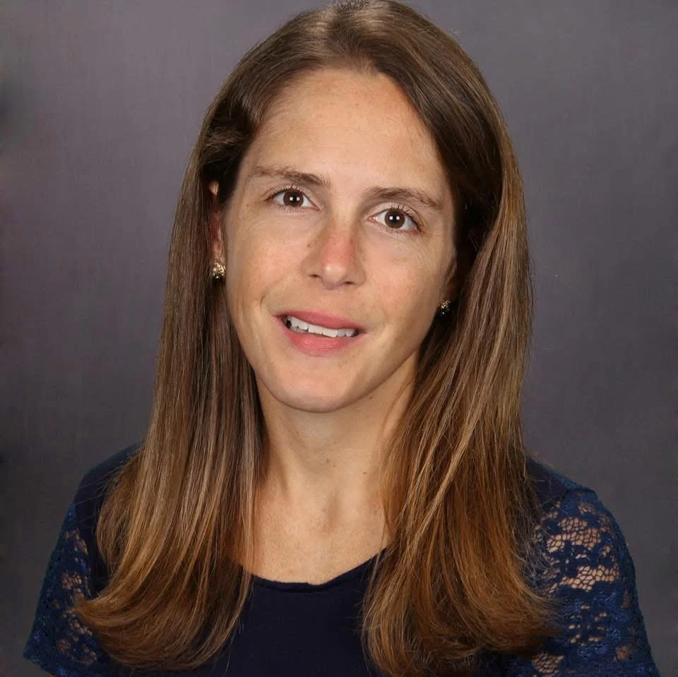 A woman with long brown hair, wearing a navy blue top with lace sleeves, smiling softly against a dark grey background.