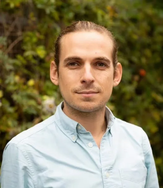 Joe Pitts-Cunnngham - a white man with brown hair tied back, wearing a light blue button-up shirt, standing outdoors in front of green foliage.