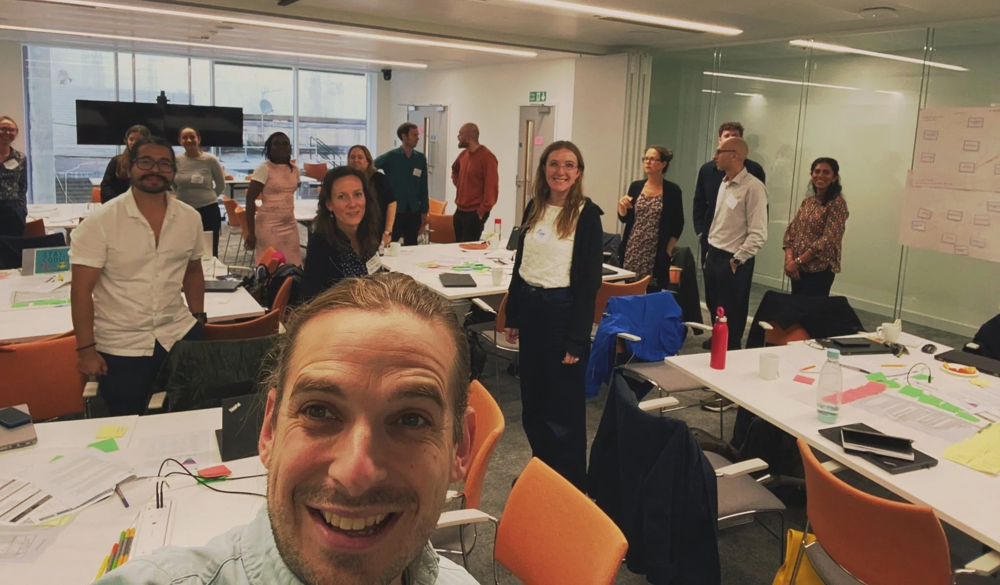 Group of diverse people gathered in a well-lit conference room, some smiling at the camera, others engaged in conversation or looking at the camera, with tables, papers, and colorful sticky notes scattered around.