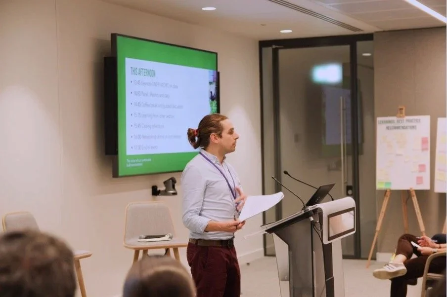 Joe Pitts-Cuninningham giving a presentation in a conference room with a green slide on the screen and an audience seated facing him.